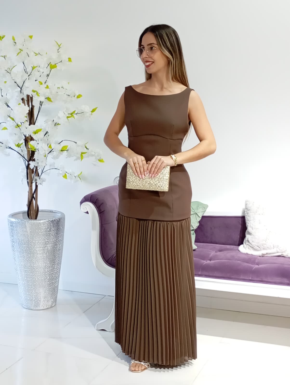 Vestido Vera&Lucy Mulher com vestido castanho e clutch dourada em interior com sofá roxo e vaso com flores