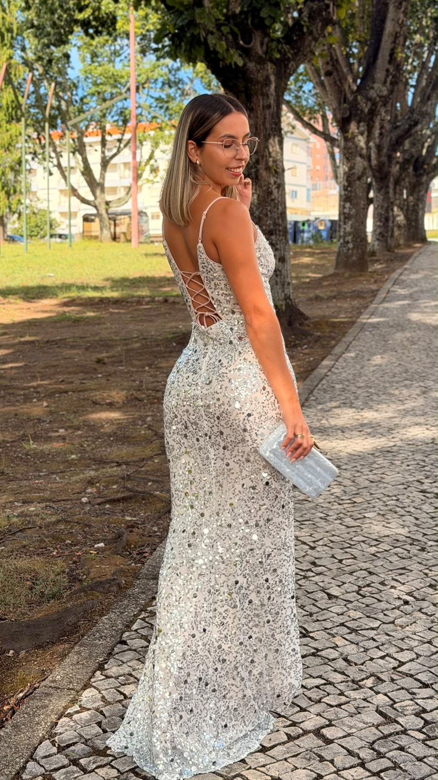 Vestido Milene Lantejoulas Mulher de costas com vestido prateado de lantejoulas e clutch azul claro ao ar livre