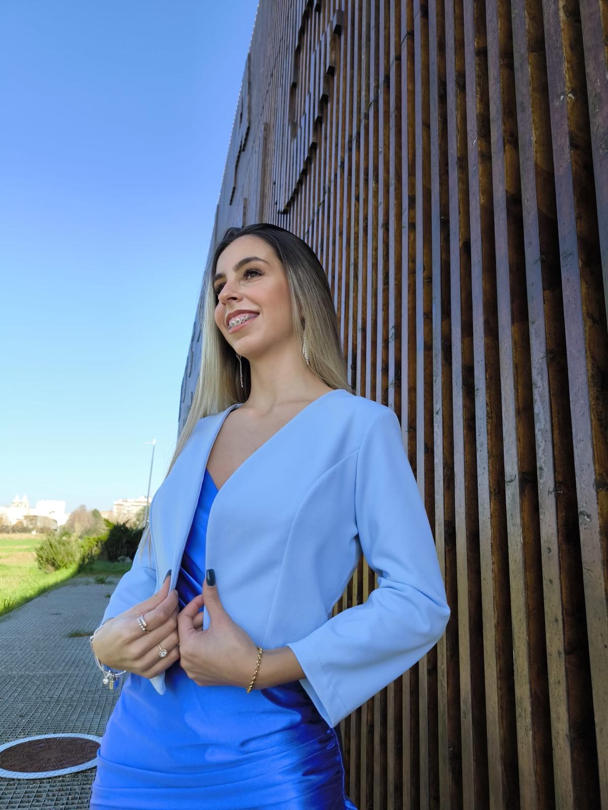 Bolero Simply Mulher com casaco azul claro e vestido azul brilhante ao ar livre junto a parede de madeira