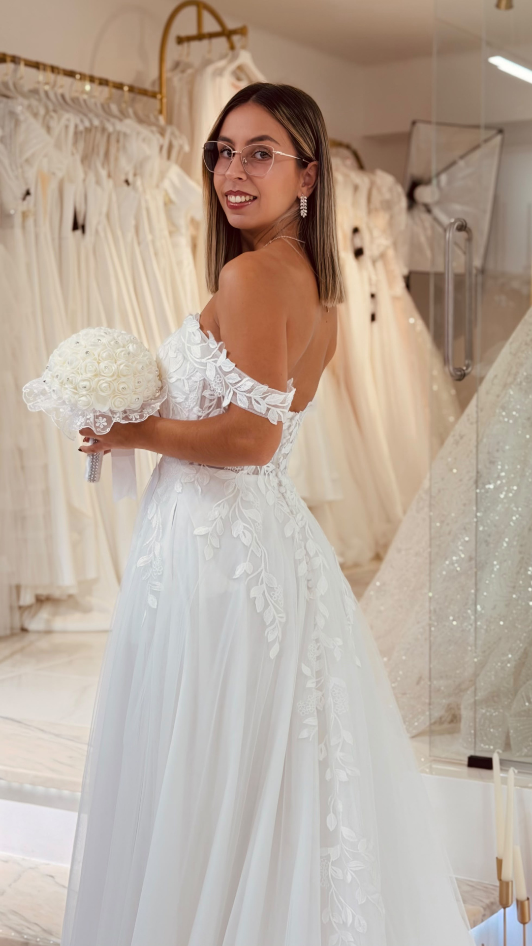 Vestido Amarílis Mulher de costas a usar vestido de noiva branco com renda e a segurar bouquet de flores brancas na loja de vestidos
