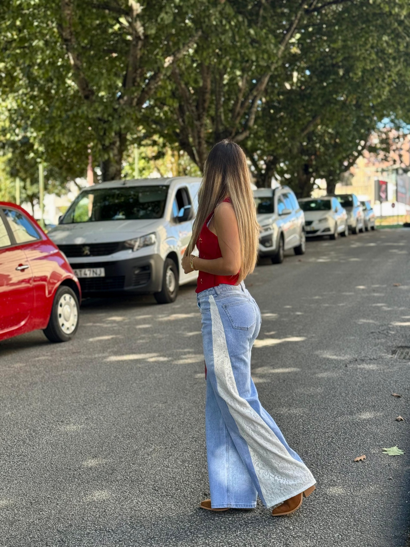 Calça de Ganga c/ Renda Branca Mulher de costas com calças de ganga bicolores e blusa vermelha numa rua com carros e árvores