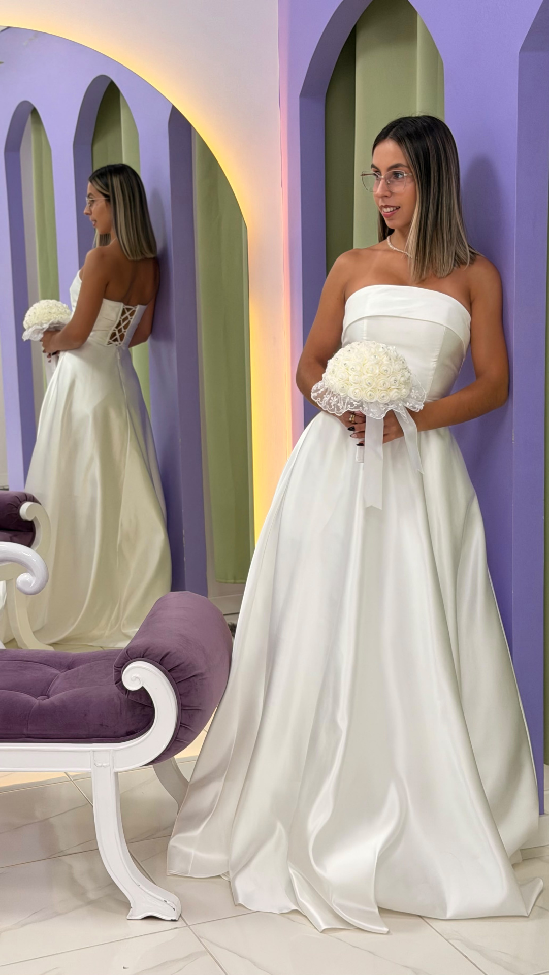 Vestido Alizee Noiva Mulher de vestido de noiva branco segurando bouquet, encostada em parede roxa com espelho e sofá roxo.
