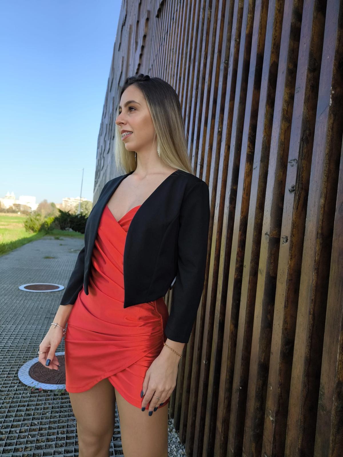 Bolero Simply Mulher com vestido vermelho justo e casaco curto preto ao ar livre junto a parede de madeira