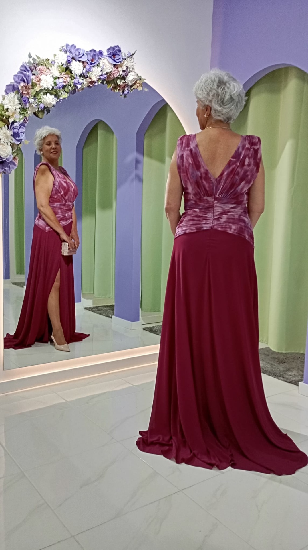 Vestido Andore Plus Size Vestido vinho com padrão tie-dye roxo e fenda, refletido num espelho com moldura de flores.