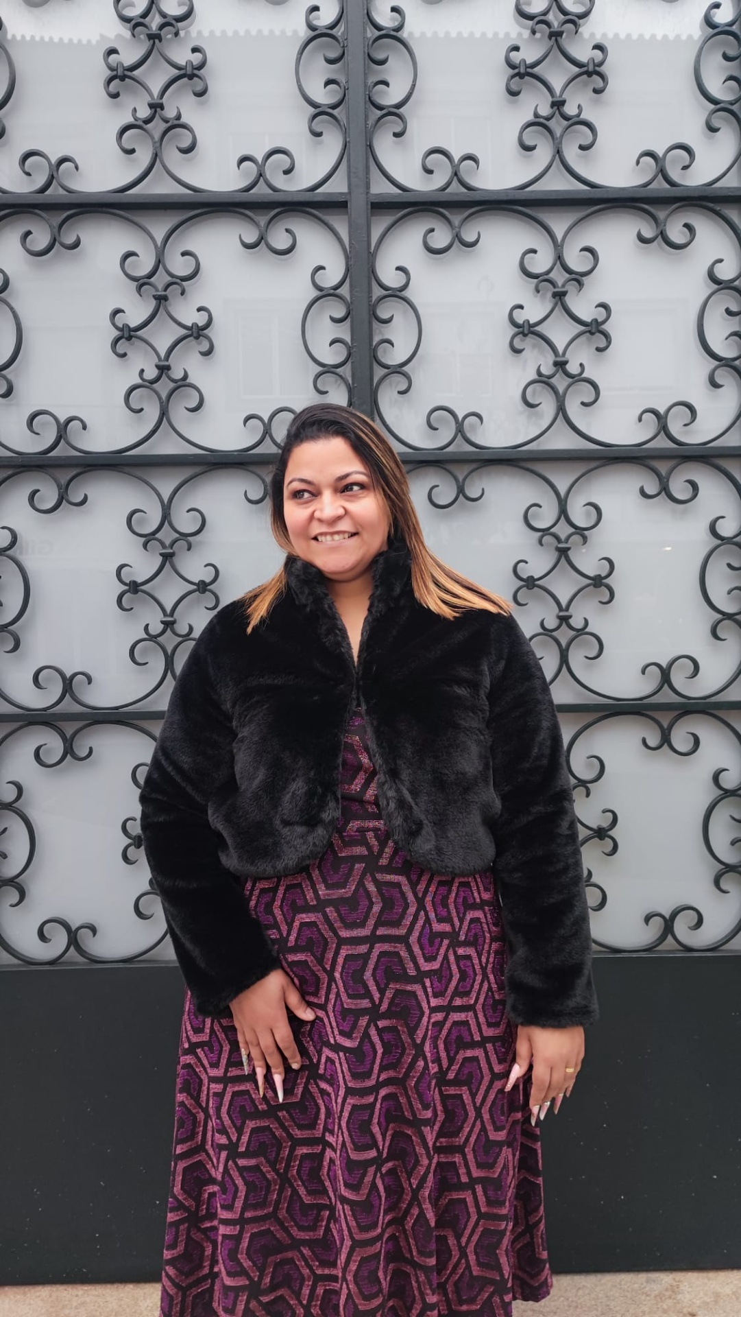 Bolero de pêlo Mulher com casaco preto de pelos curtos e vestido geométrico roxo em frente a uma parede com grades de ferro decorativas pretas