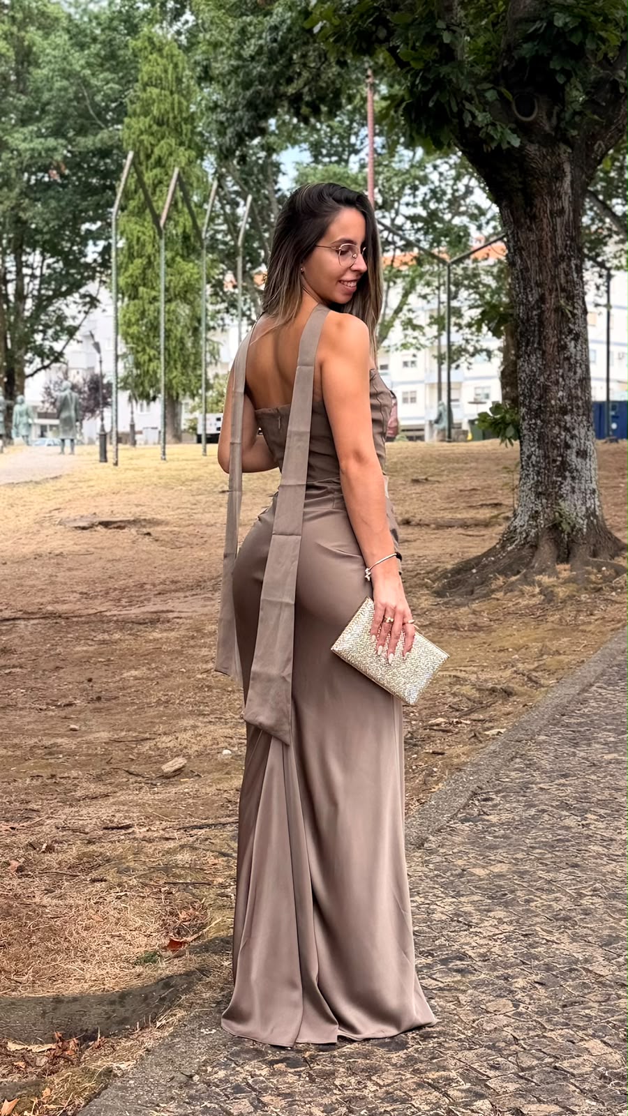 Vestido Romana Mulher com vestido castanho longo e alças largas segurando carteira prateada em parque com árvores