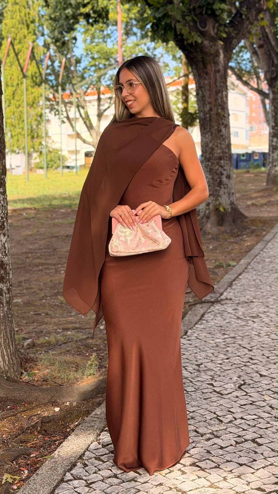Vestido F&P MIRAGE Castanho Mulher com vestido castanho longo e capa, mala rosa bordada e acessórios dourados ao ar livre