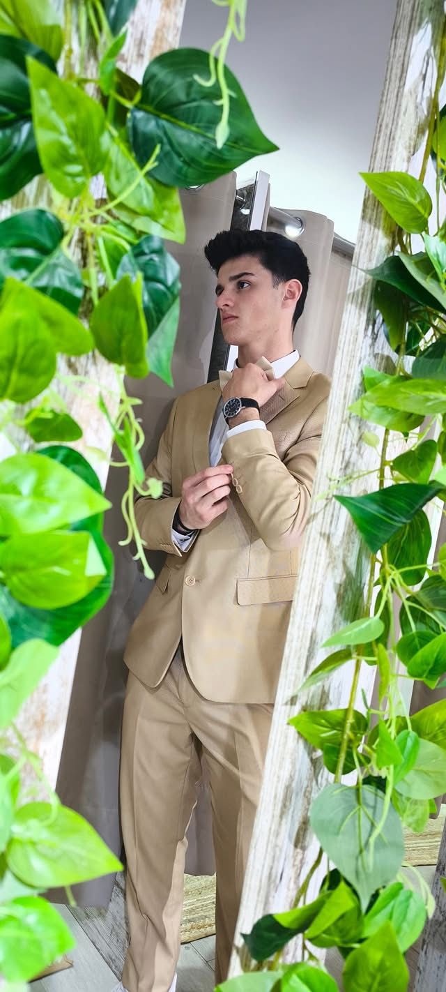 Fato Gilgavin Bege Ponteado Homem com fato bege e camisa branca ajustando a gola, com folhas verdes ao redor