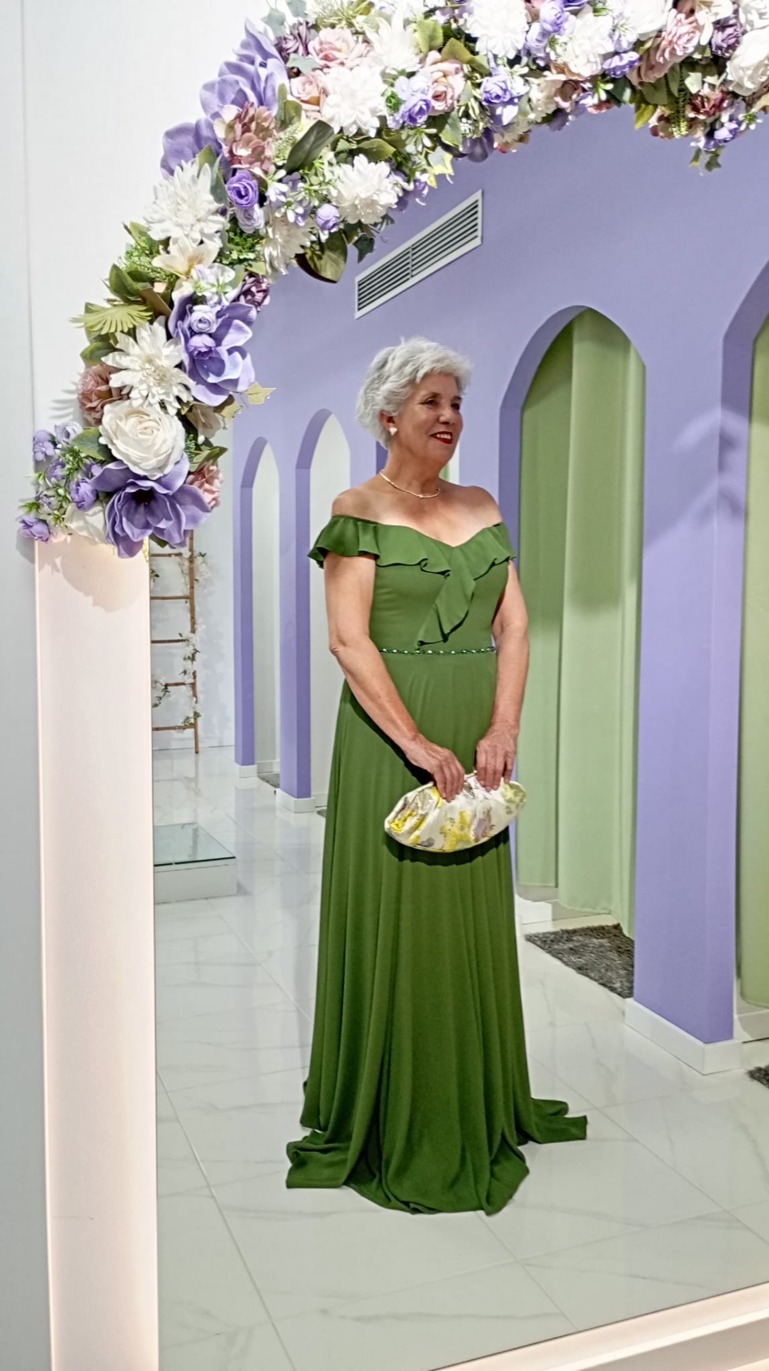 Vestido Nataly Mulher com vestido verde longo e bolsa branca e amarela em ambiente com parede lilás e arco de flores.