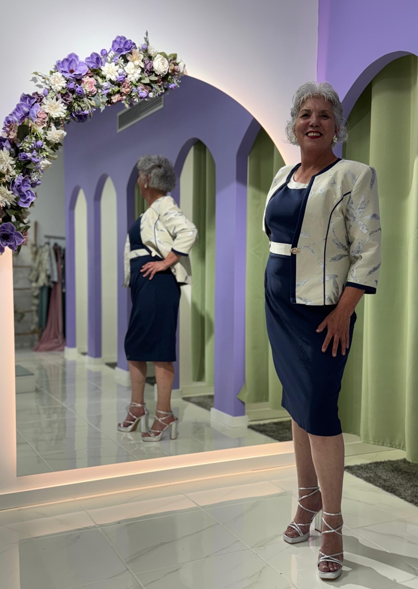 Conj. Casaco + Vestido Serena Mulher com vestido azul escuro e casaco bege com padrão floral junto a espelho com decoração floral roxa.