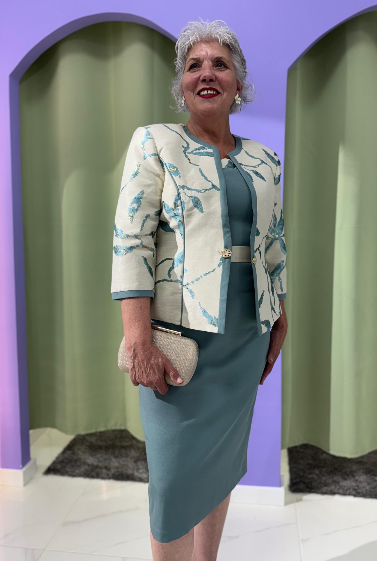 Conj. Casaco + Vestido Serena Conjunto feminino vestido azul-petróleo e casaco creme com padrão azul em ambiente interior.