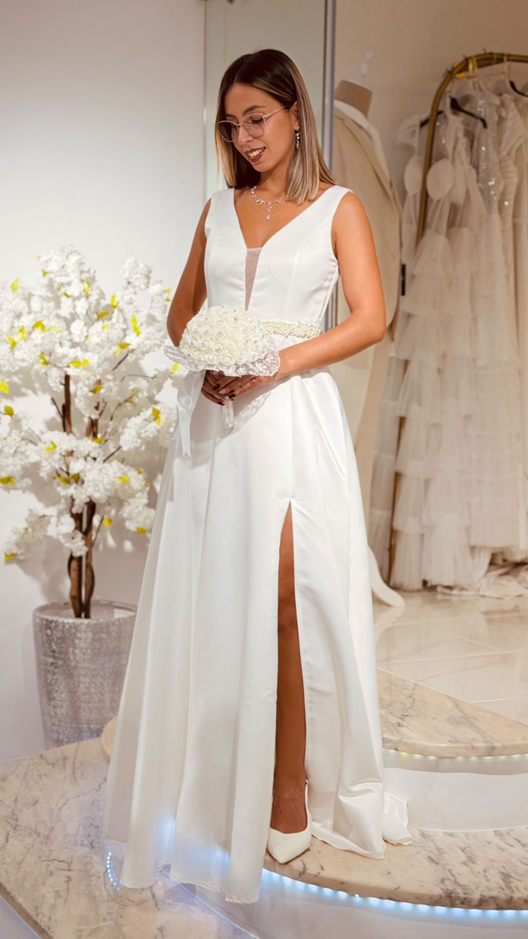 Vestido Glace Noiva Vestido de noiva branco com decote em V, cinto de pérolas e fenda lateral, bouquet de rosas brancas, mulher com óculos e acessórios prateados.