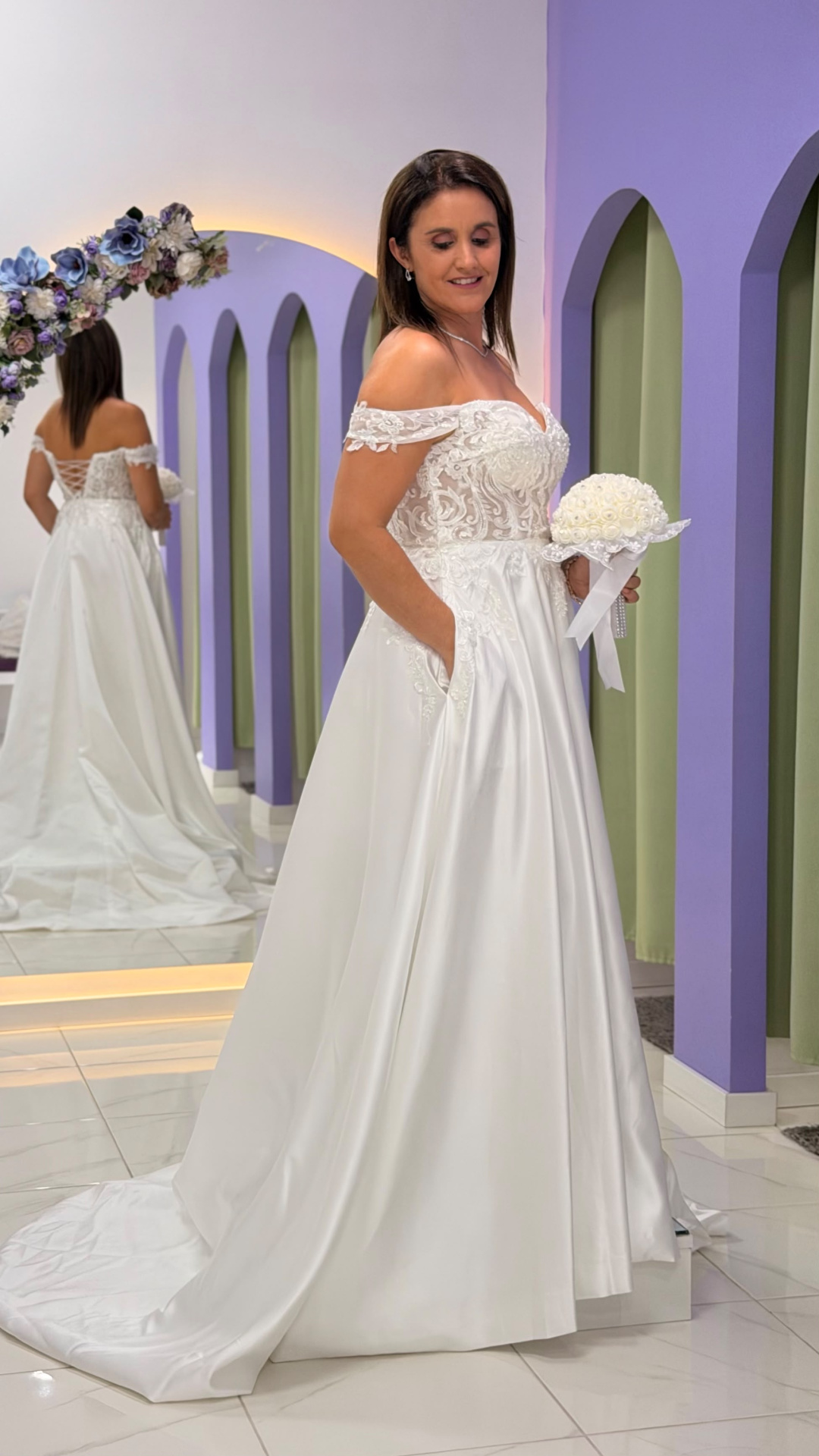 Vestido Amour Glamour Mulher com vestido de noiva branco com detalhes em renda, segurando bouquet branco, em frente a um espelho num ambiente com paredes lilás e cortinas verdes