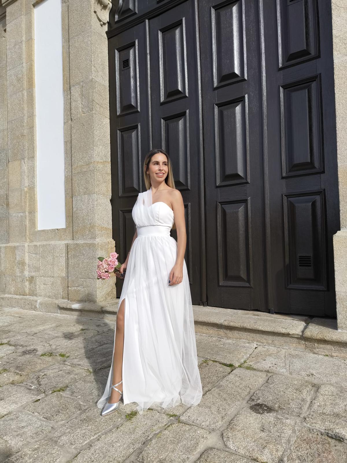 Elegância Minimalista - Vestido Noiva Mulher com vestido branco longo com fenda, segurando buquê de flores cor-de-rosa, em frente a porta de madeira escura.