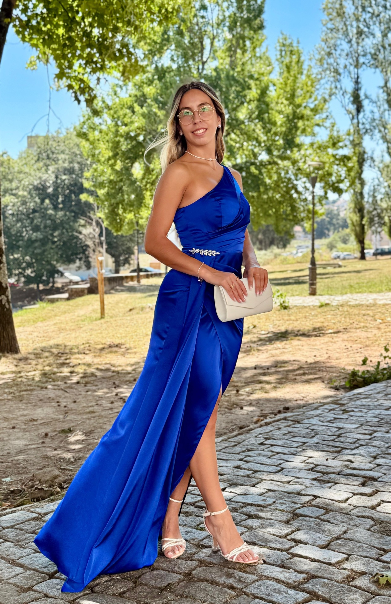Vestido Mayse Mulher com vestido azul royal de um ombro só, sandálias brancas e mala bege ao ar livre