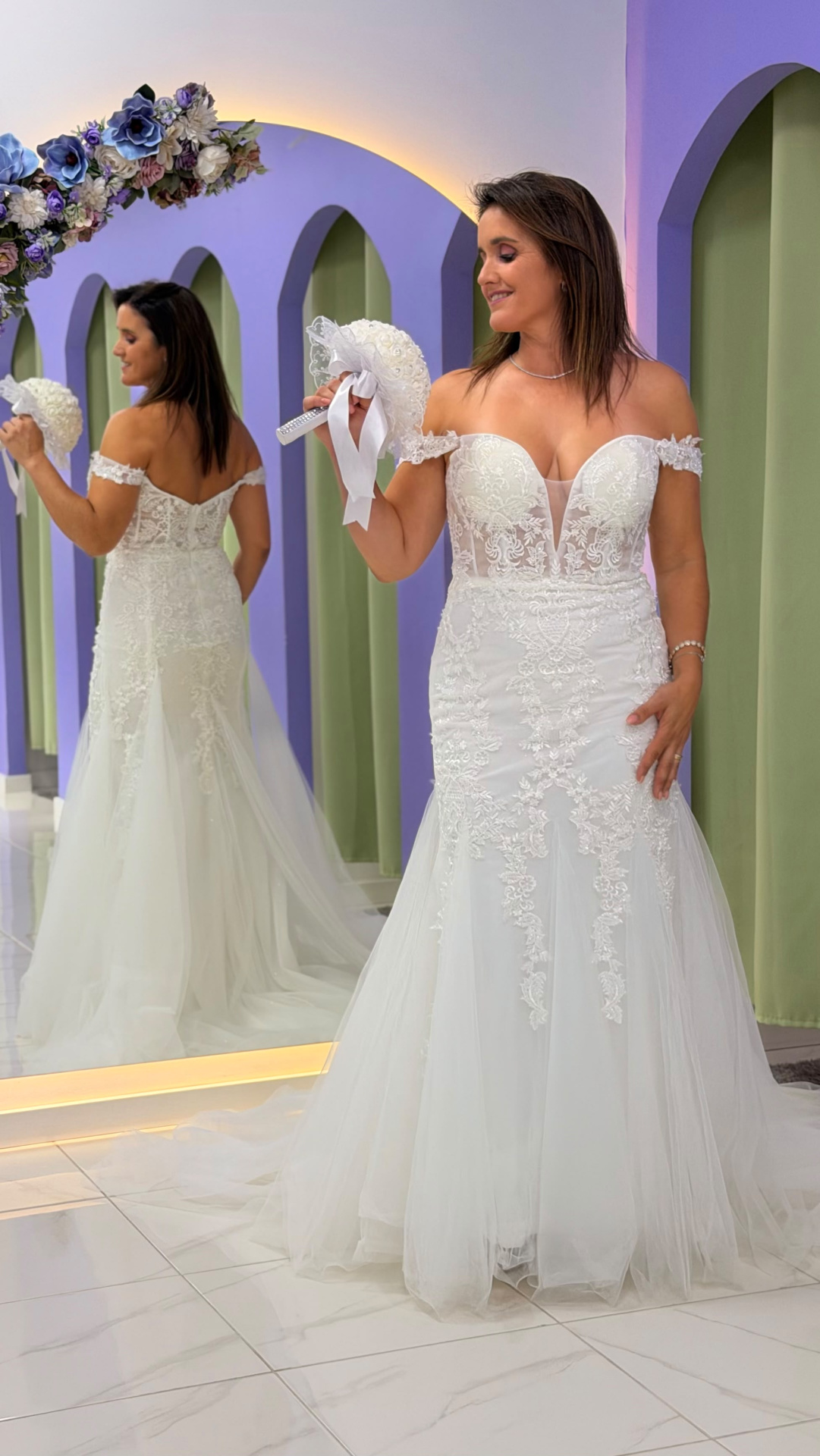 Vestido Wedding Ilusion Vestido de noiva branco com renda e tule, decote coração, segurando bouquet branco, refletido em espelho num ambiente com arcos lilás e cortinas verdes.