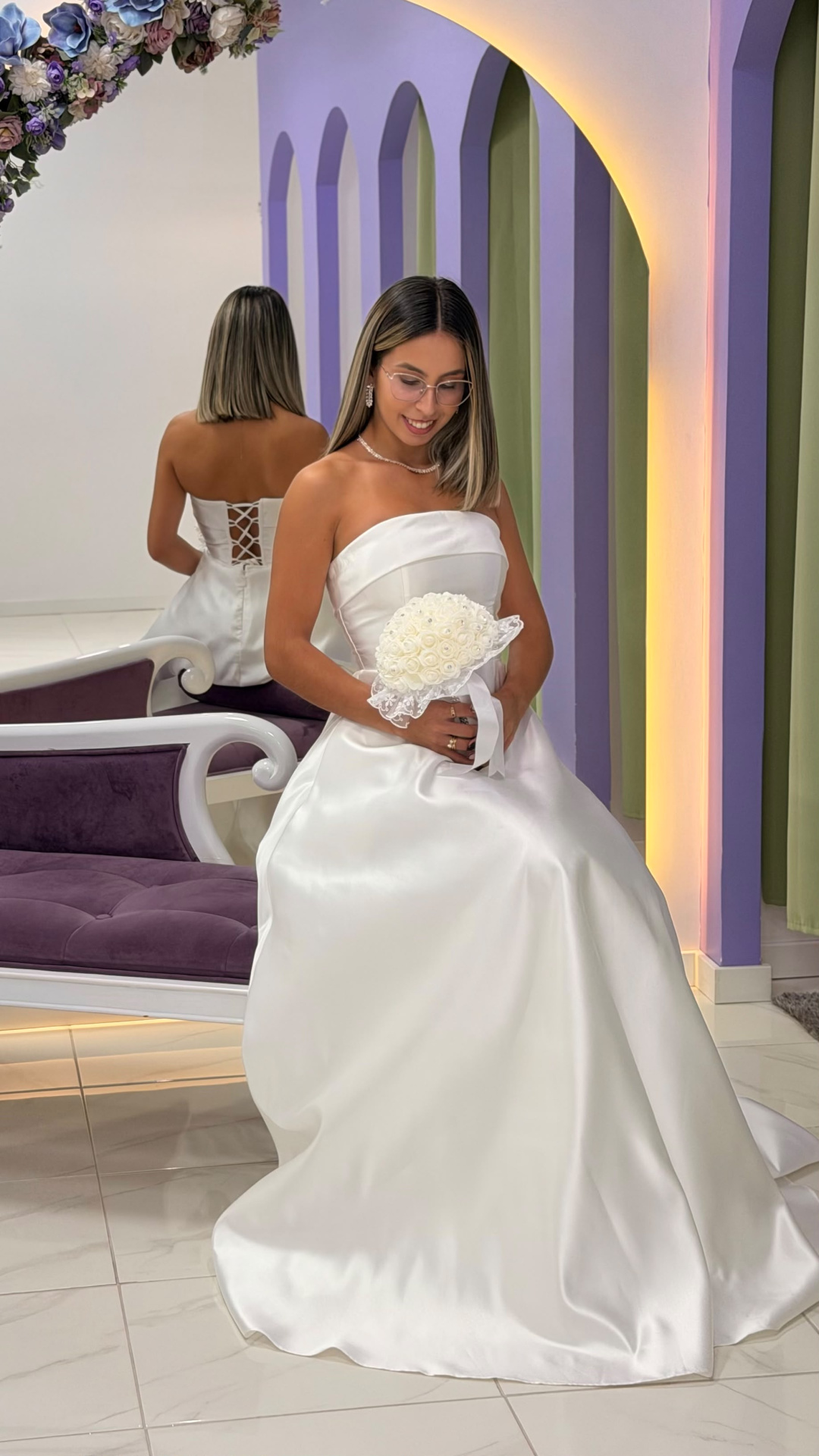 Vestido Alizee Noiva Vestido de noiva branco com bouquet e banco roxo em estúdio decorado com arco e flores.
