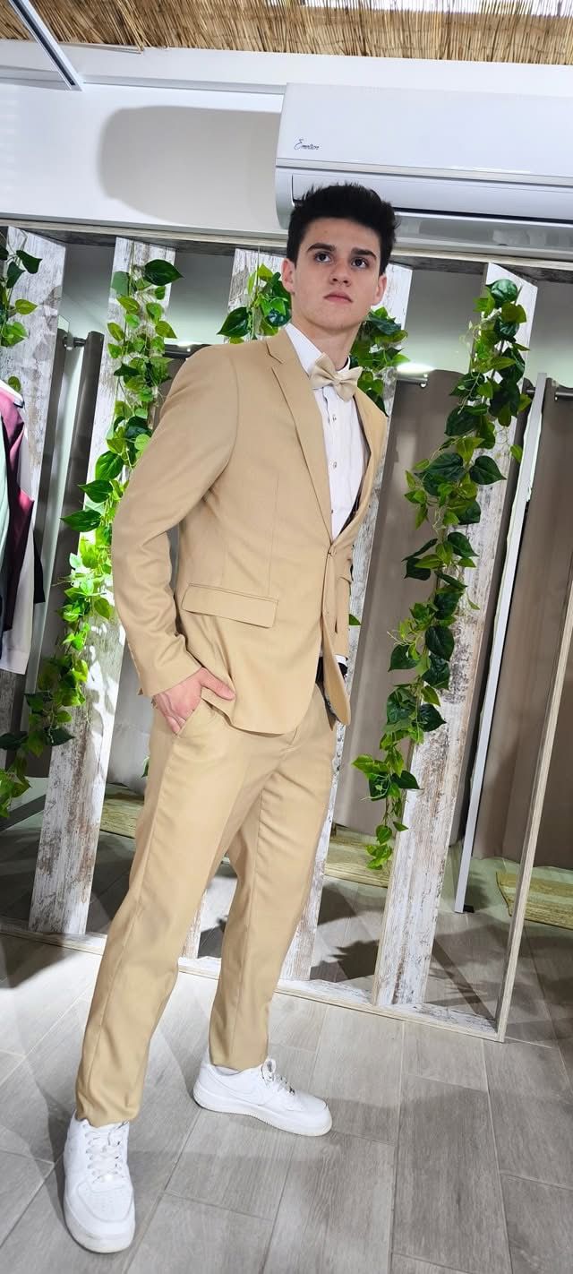 Fato Gilgavin Bege Ponteado Homem com fato bege, camisa branca, gravata borboleta bege e ténis brancos num ambiente interior com decoração de madeira e plantas.