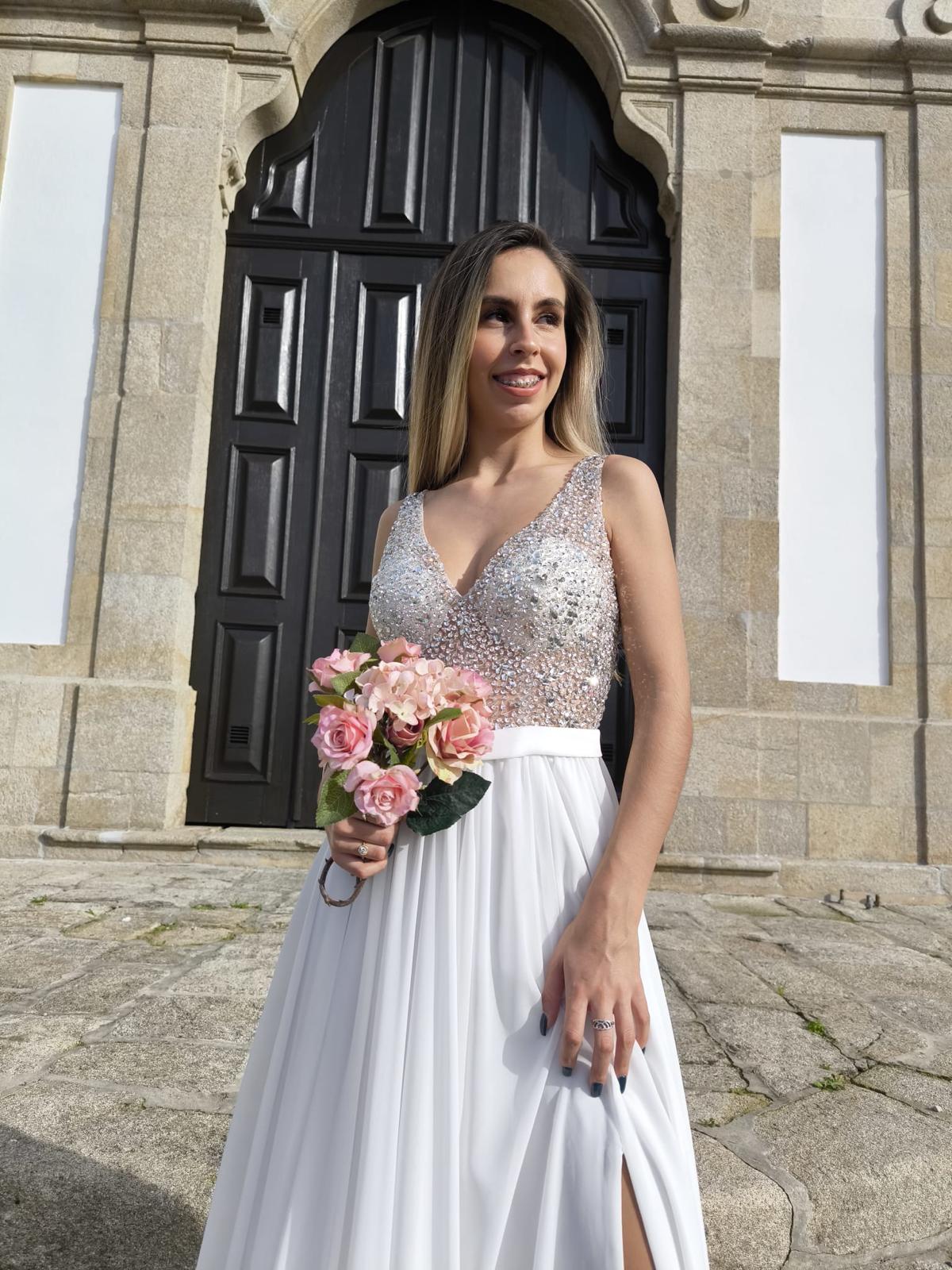 Vestido Simplicidade Serena Vestido de noiva branco com corpete de lantejoulas e ramo de flores rosas ao ar livre
