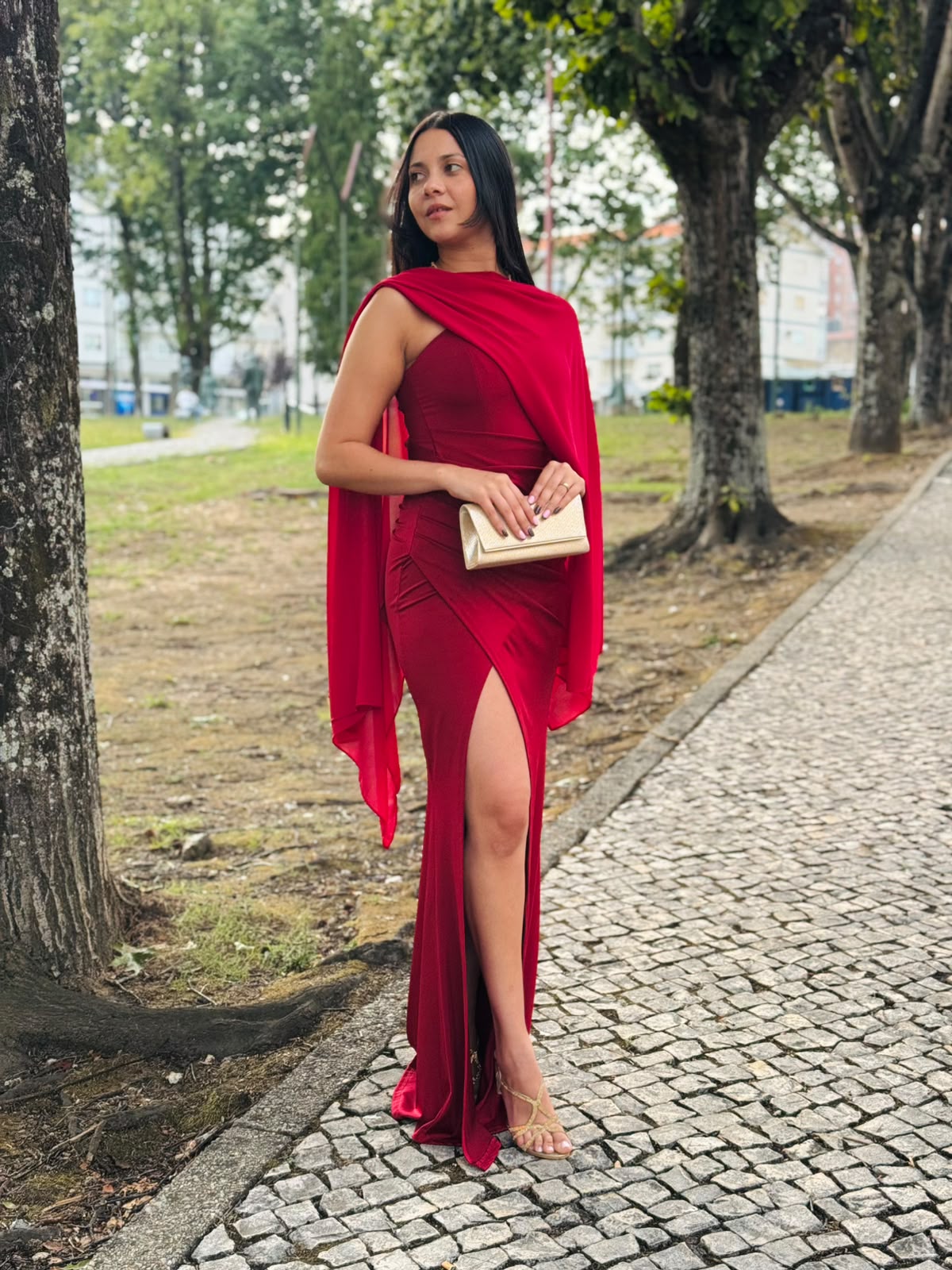 Vestido HUNGARO Mulher com vestido longo vermelho e mala clutch dourada num passeio com árvores.