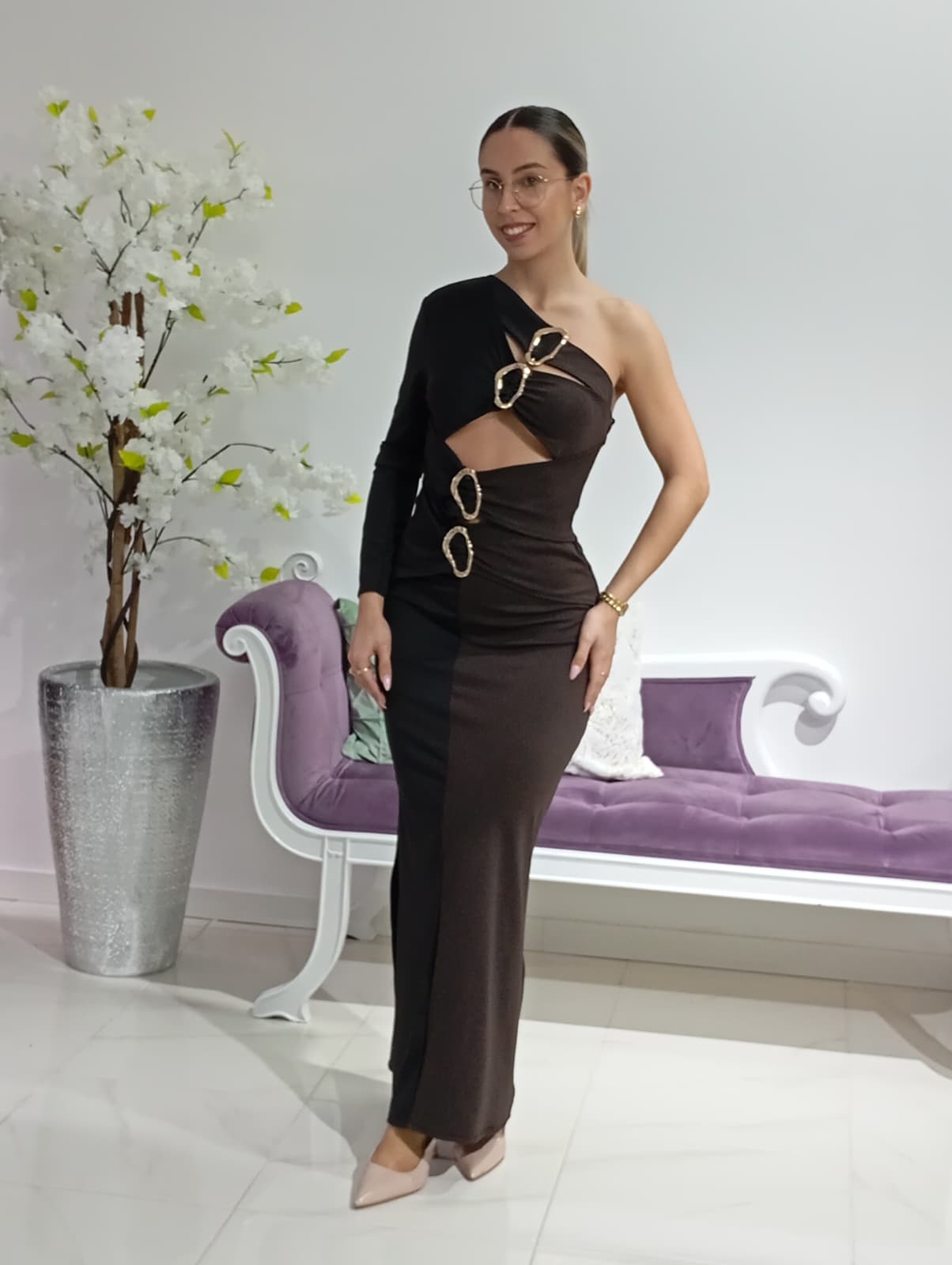 Vestido Savannah Preto/Castanho Mulher com vestido preto assimétrico com argolas douradas, sofá roxo e vaso com flores brancas