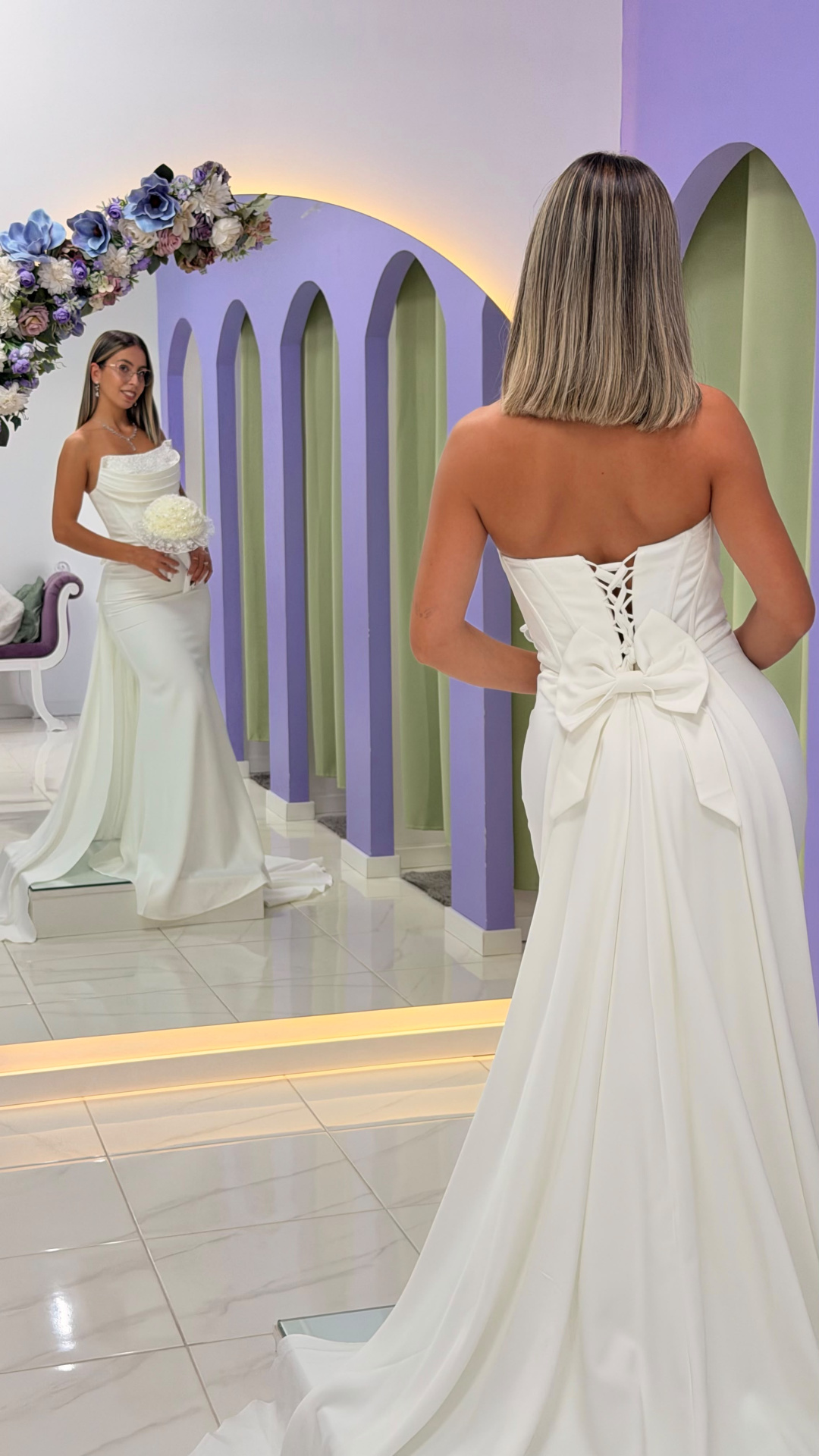 Vestido Éden Noiva Vestido de noiva branco com laço atrás refletido num espelho num ambiente com arcos lilás