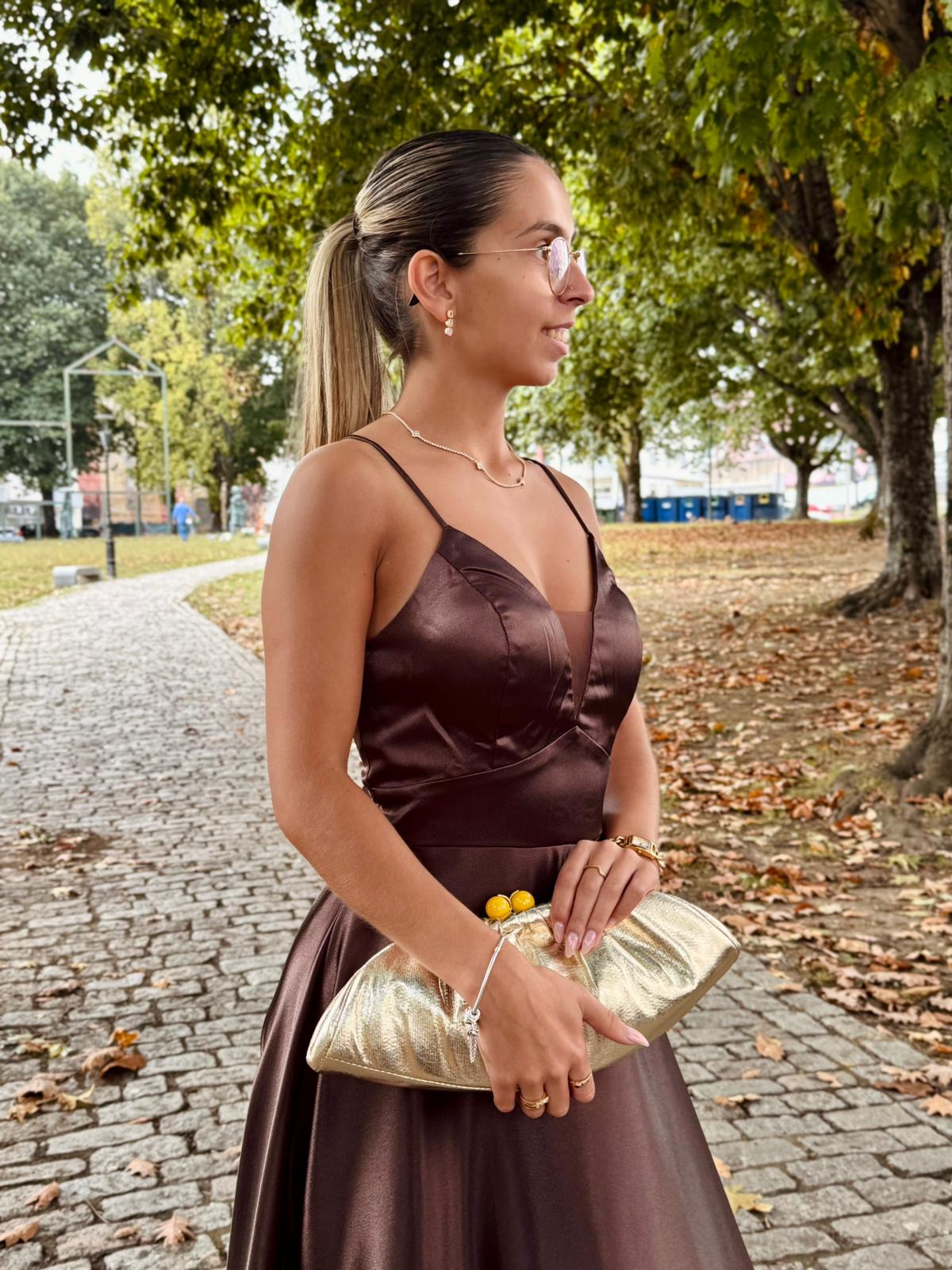 Vestido Doriane Mulher de vestido castanho acetinado e mala dourada numa rua de pedra com árvores