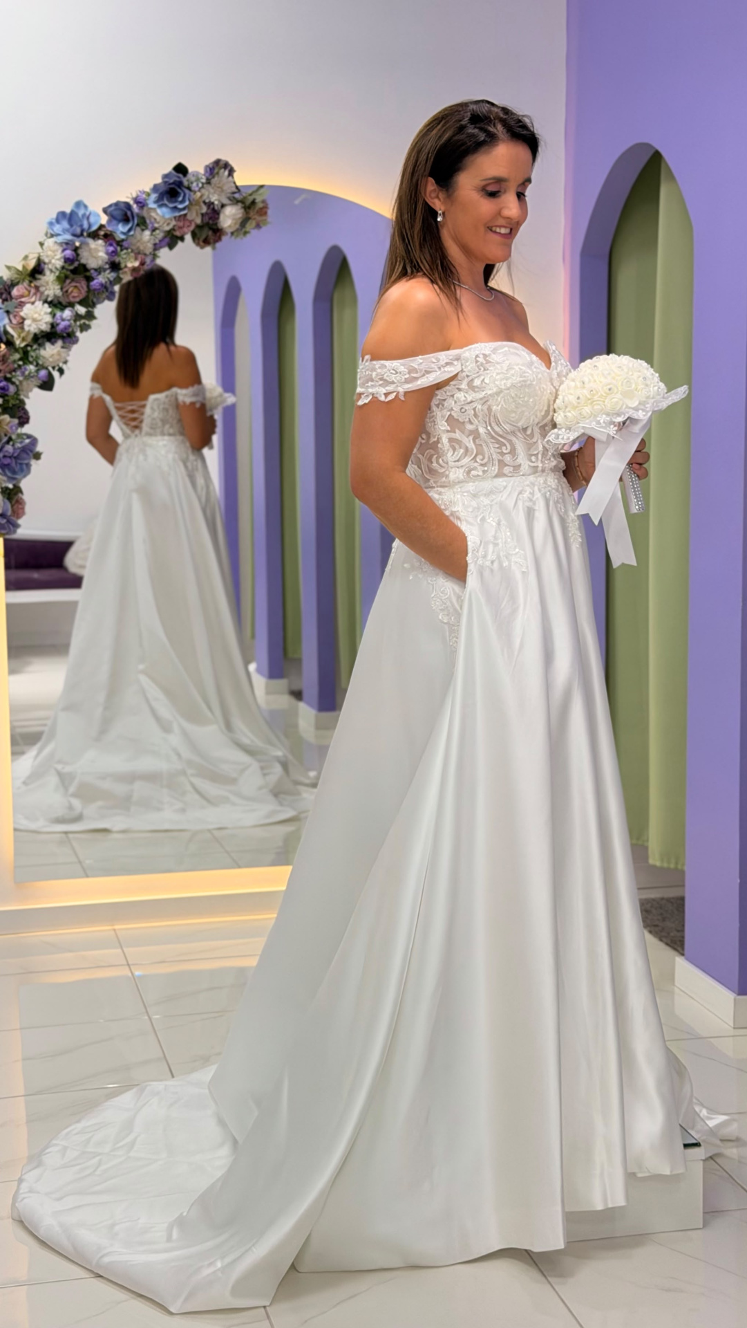 Vestido Amour Glamour Mulher com vestido de noiva branco de renda e cetim, segurando ramo, em loja com decoração roxa e espelho.