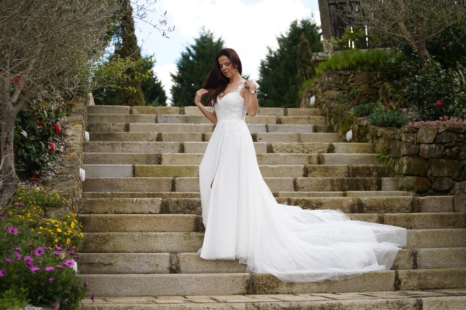 Vestido Amarílis Vestido de casamento branco com renda e flores, com saia longa fluída, em escadaria de pedra exterior.