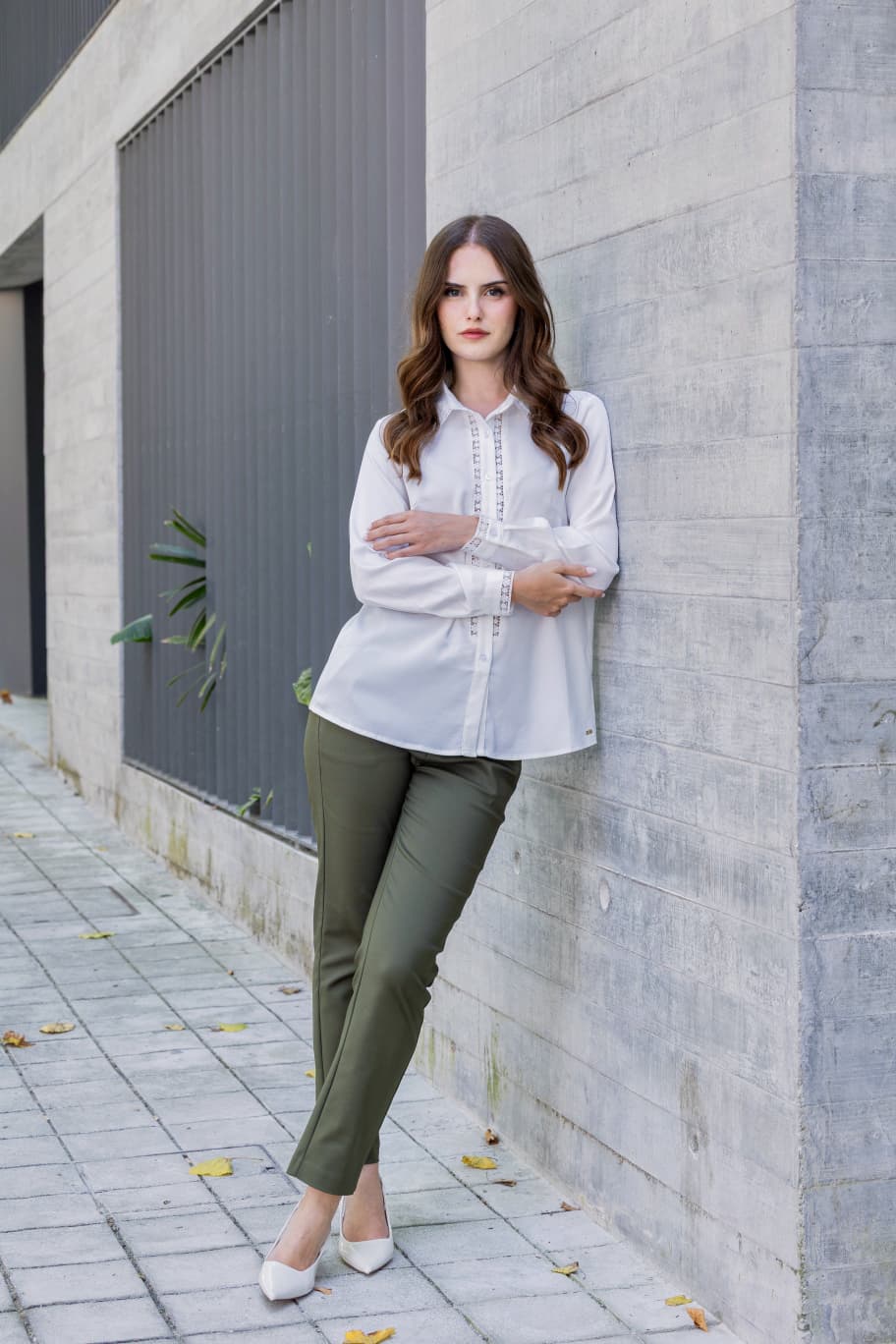 Blusa c/renda Moda Ana Mulher vestindo camisa branca com detalhes em renda e calças verdes, encostada numa parede de betão