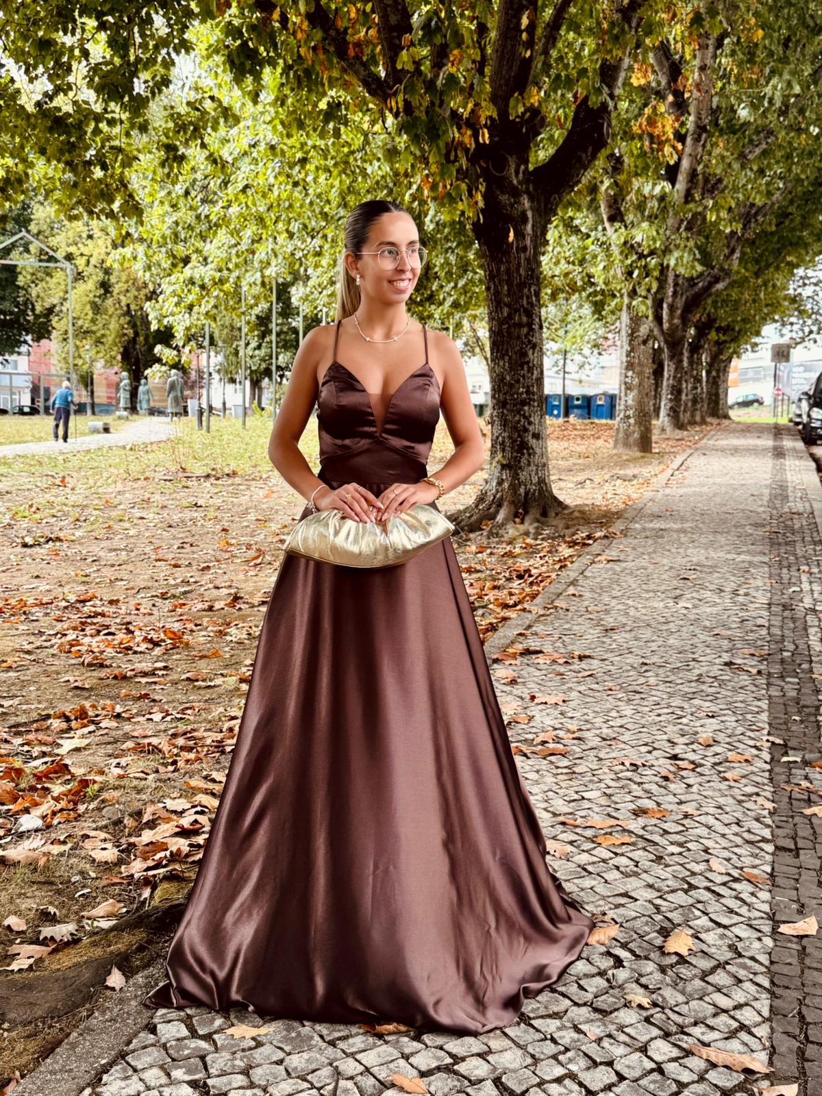 Vestido Doriane Mulher com vestido castanho acetinado segurando mala de mão creme em passeio com árvores