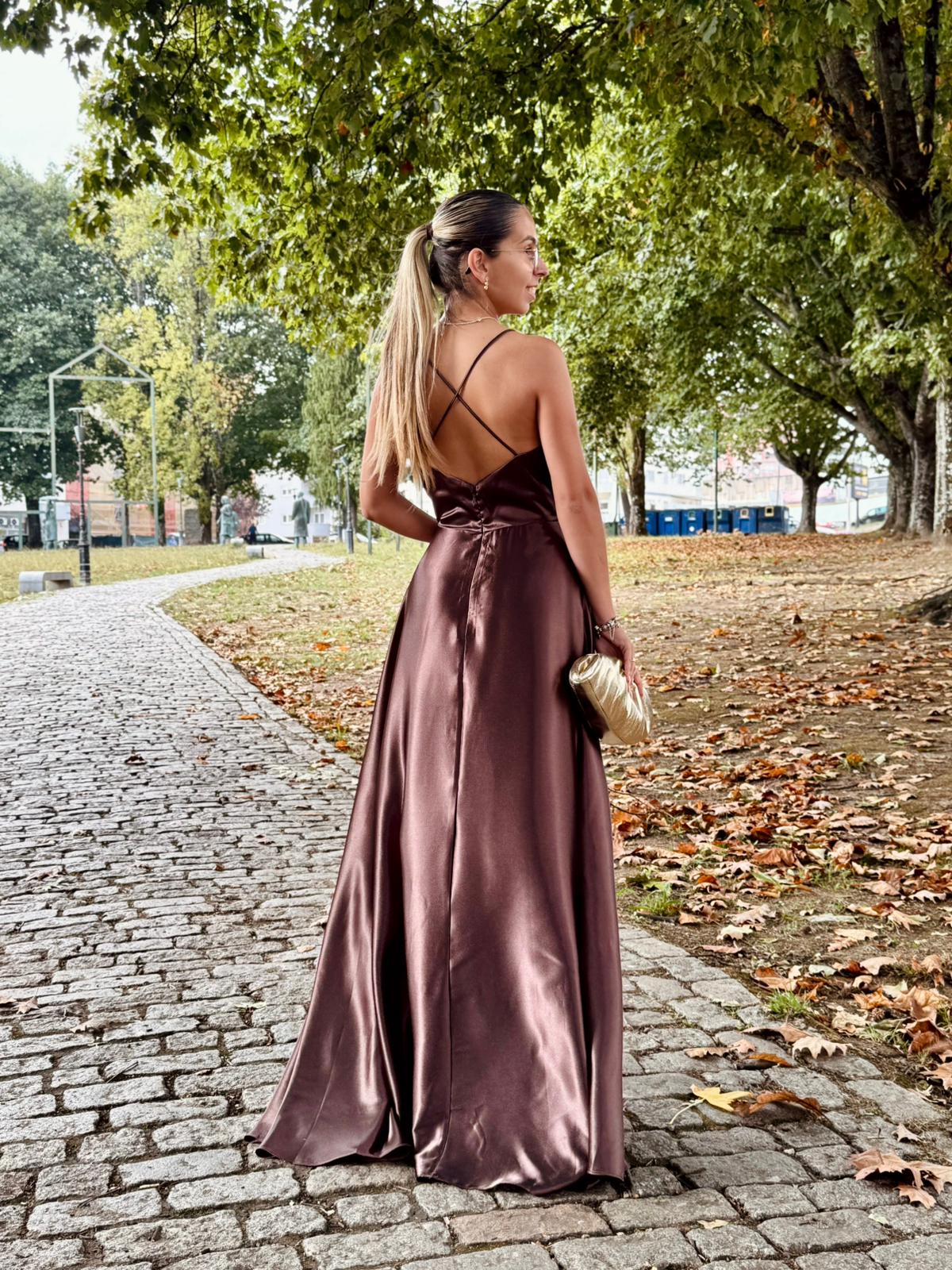 Vestido Doriane Vestido longo acetinado castanho chocolate com alças cruzadas e mala pequena dourada em passeio ao ar livre