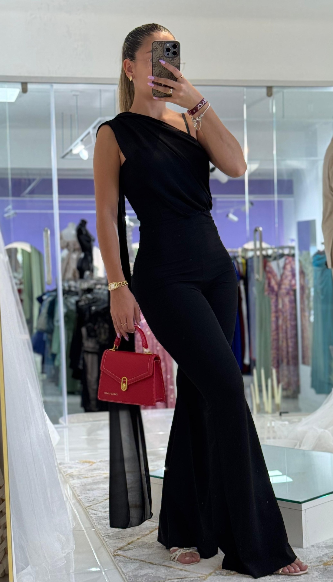 Macacão ERK c/ Echarpe Mulher com macacão preto e mala vermelha numa loja de roupas com espelhos