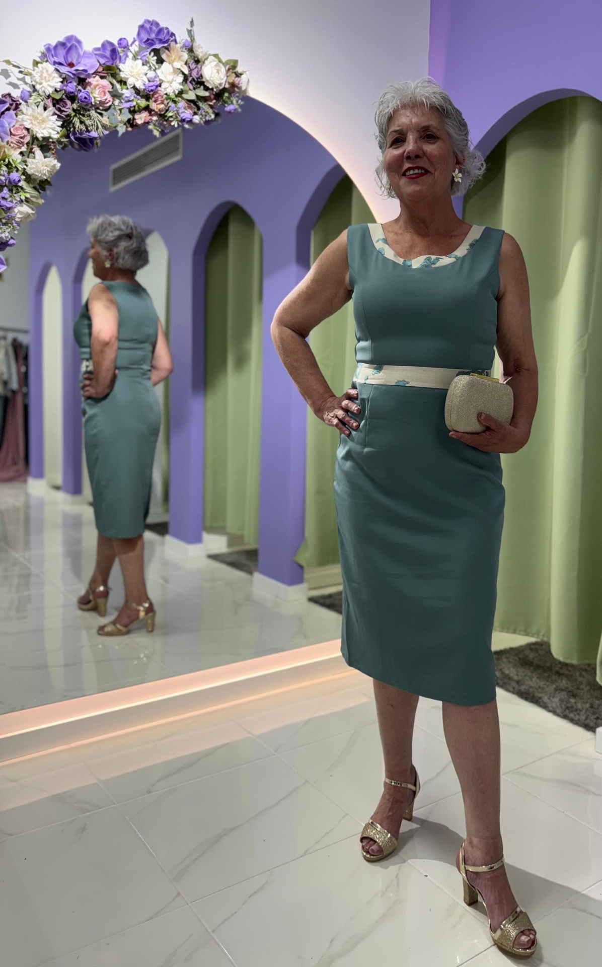 Conj. Casaco + Vestido Serena Mulher num vestido azul esverdeado com detalhes bege, sapatos dourados e mala bege, em loja com parede roxa e espelho.