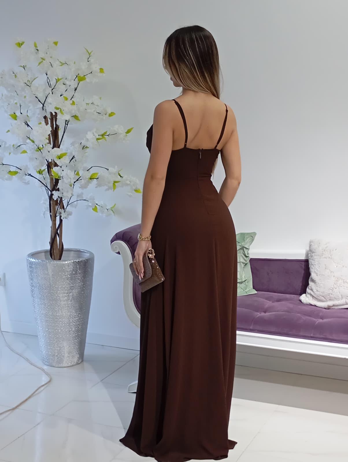 Vestido Nova Zelandia Castanho Mulher de costas com vestido longo castanho e acessório de mão sentado num ambiente interior moderno.
