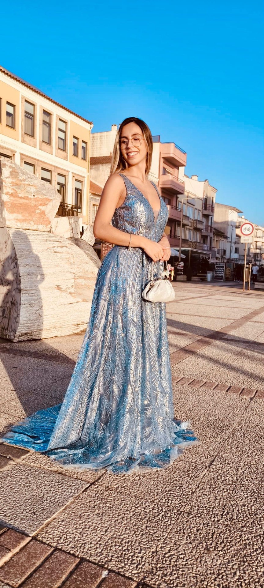 Vestido Camila Glow Mulher com vestido azul claro comprido e brilhante numa rua com edifícios.