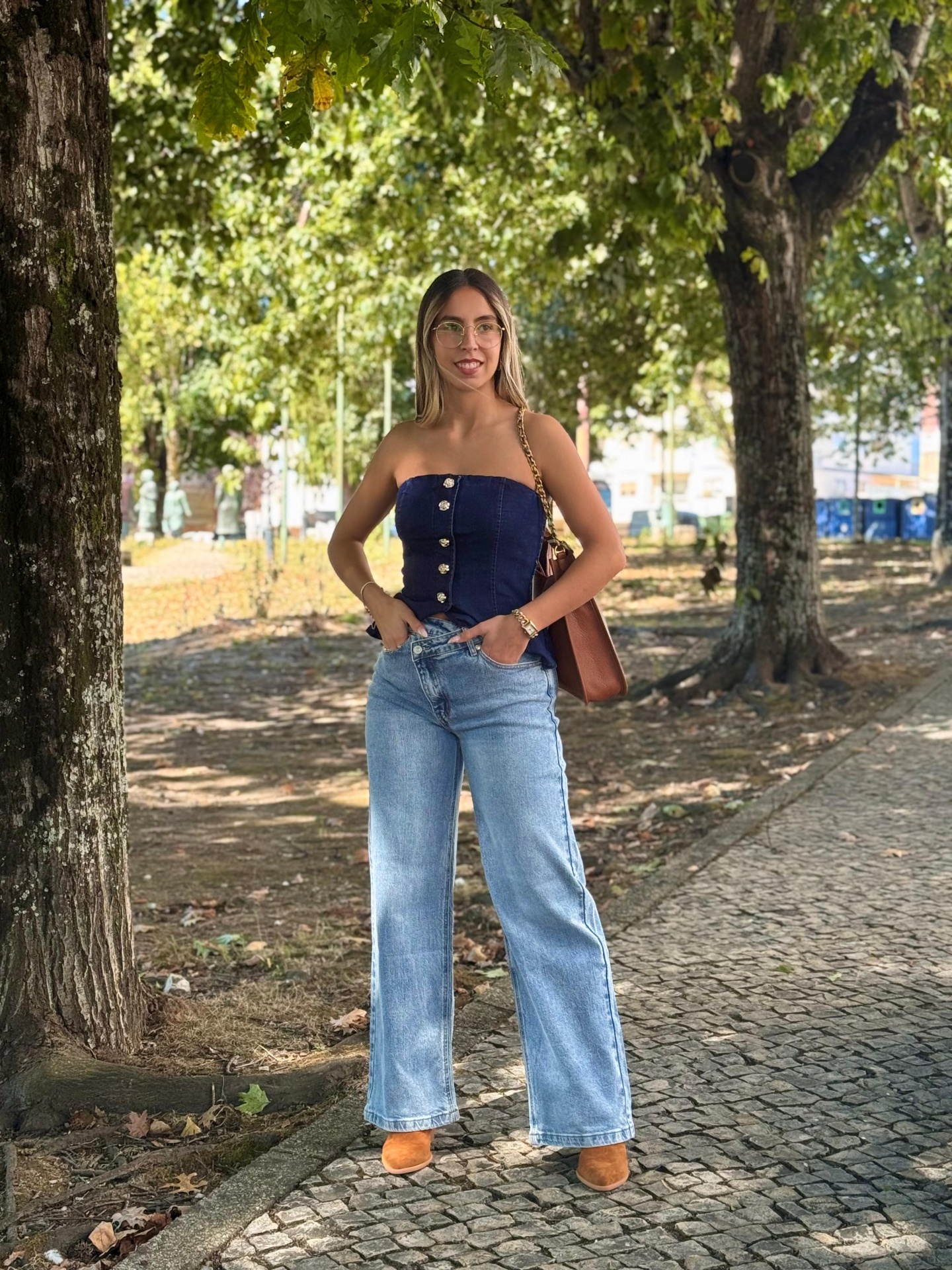 Calça de Ganga Foly Mulher jovem com top azul, calças jeans largas e bolsa castanha num parque com árvores.