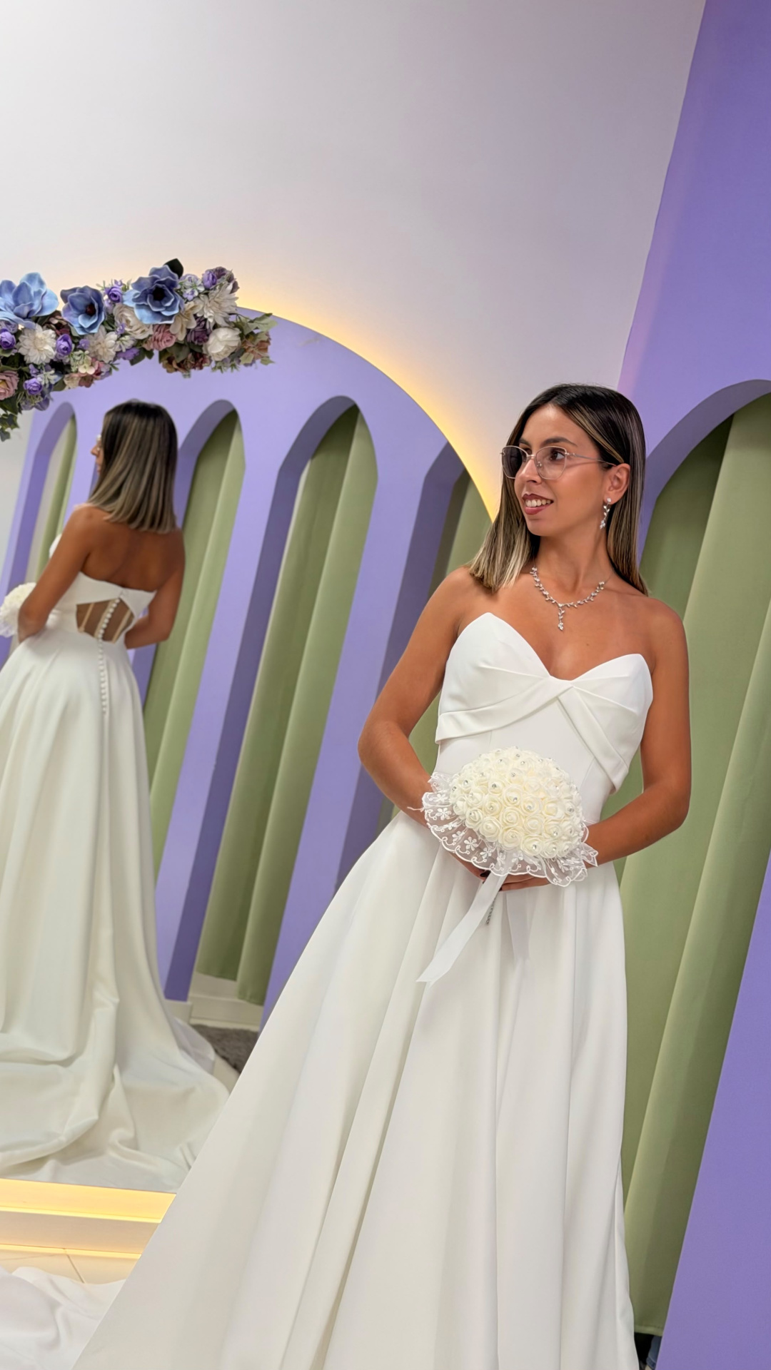 Vestido Celine Noiva Mulher com vestido de noiva branco, óculos e bouquet em frente a espelho com decoração floral