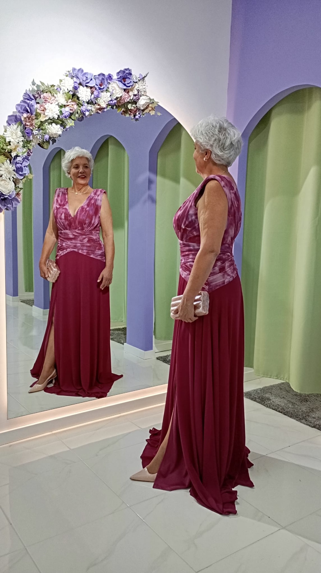 Vestido Andore Plus Size Mulher com vestido longo bordeaux com padrão tie-dye em frente a espelho decorado com flores roxas e brancas