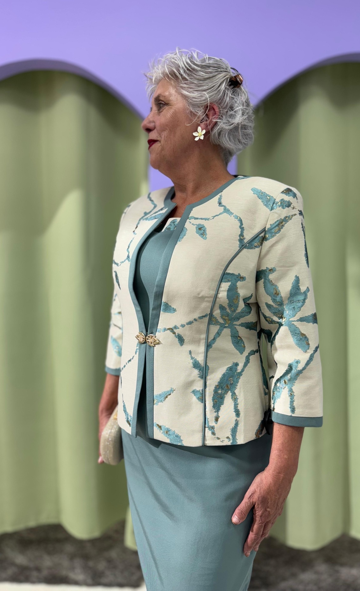 Conj. Casaco + Vestido Serena Mulher com casaco estampado de folhas azuis e vestido azul-petróleo em fundo de cortinas roxas e verdes