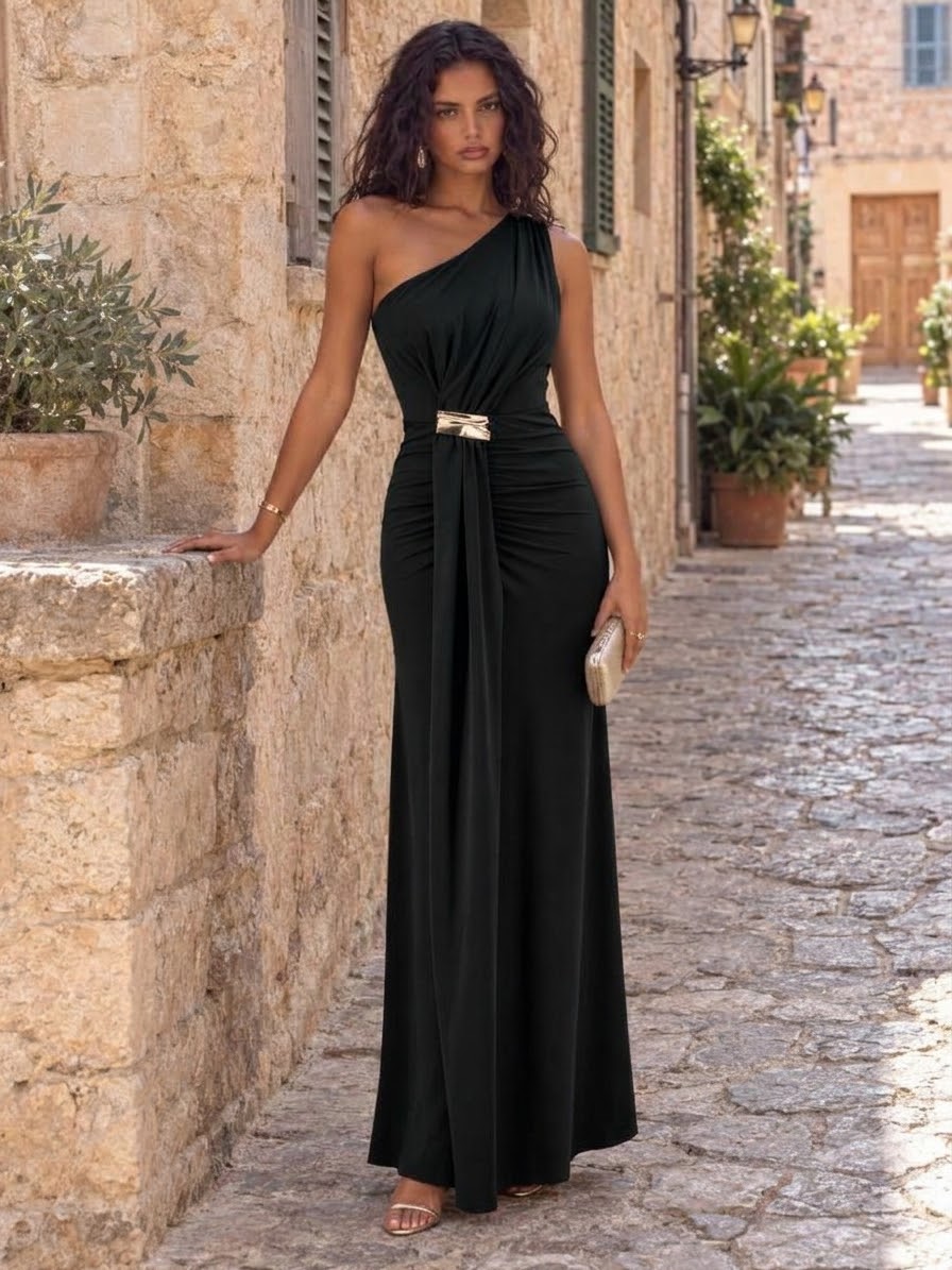 Vestido Siloé Vestido preto longo com um ombro só e faixa dourada na cintura em cenário com paredes e calçada de pedra