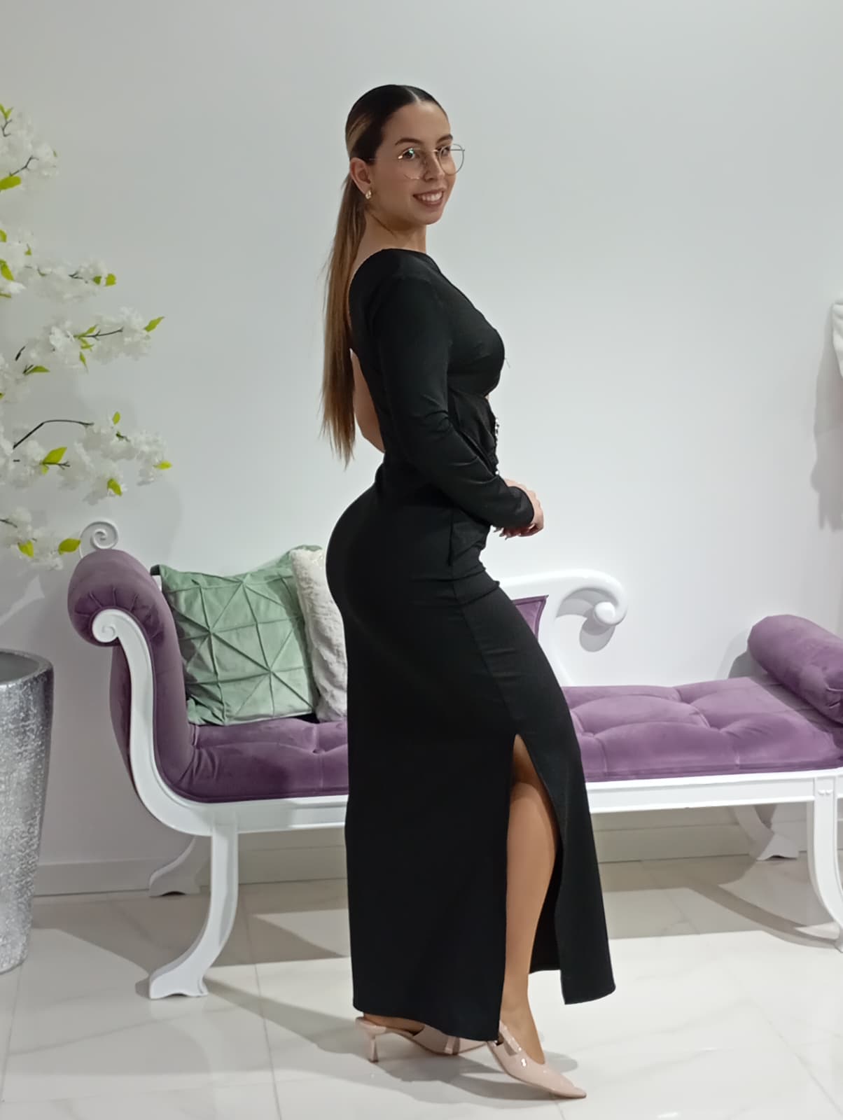 Vestido Savannah Preto/Castanho Mulher com vestido preto longo e óculos, ao lado de sofá branco com almofadas coloridas.