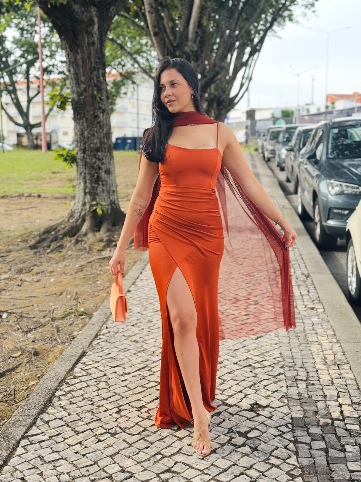 Vestido HUNGARO Mulher com vestido laranja longo com fenda e alças finas, segurando mala laranja e andando numa calçada de pedra.