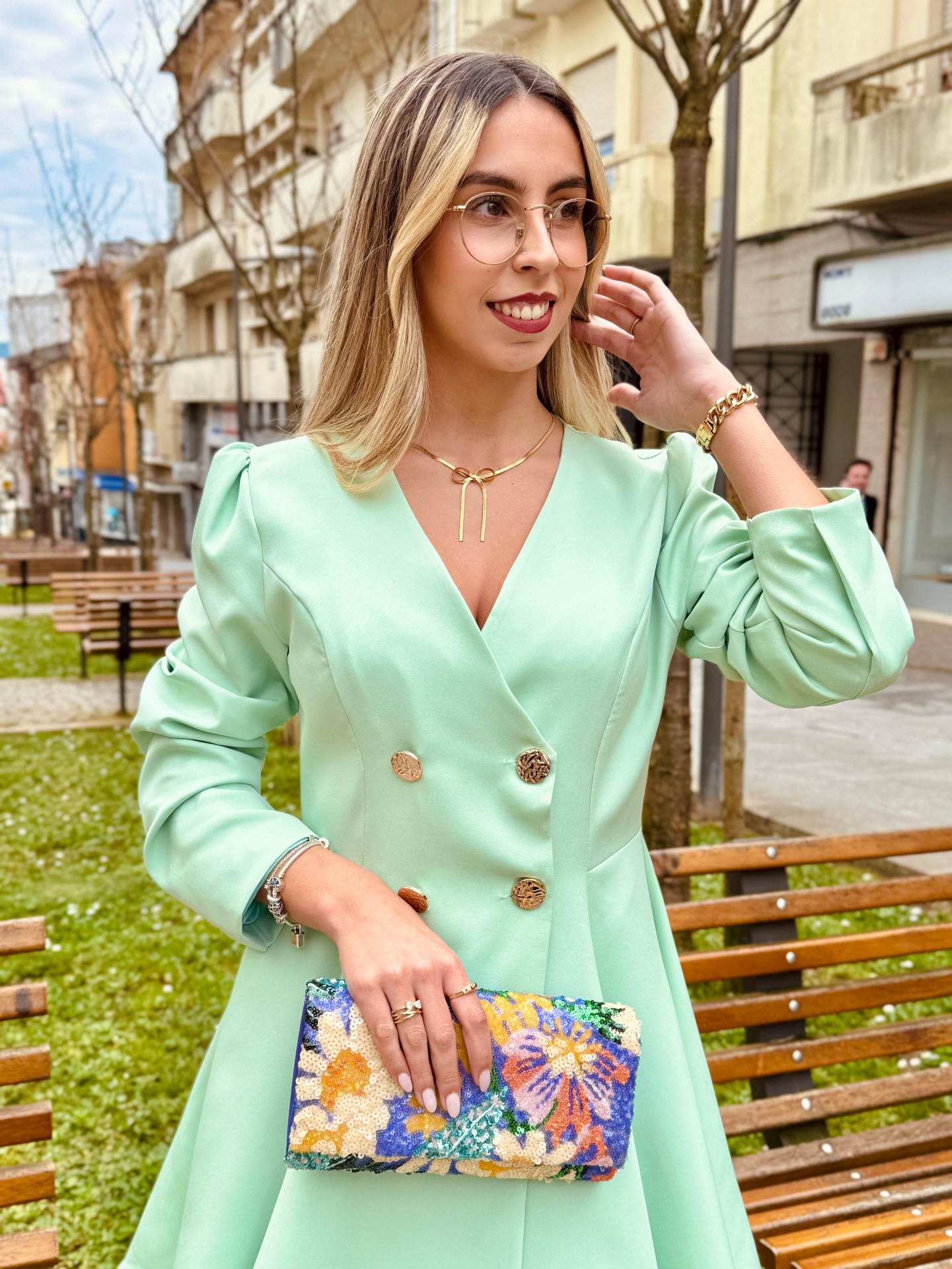 Vestido Grace Mulher vestindo vestido verde claro com botões dourados segurando mala de mão floral colorida em parque urbano
