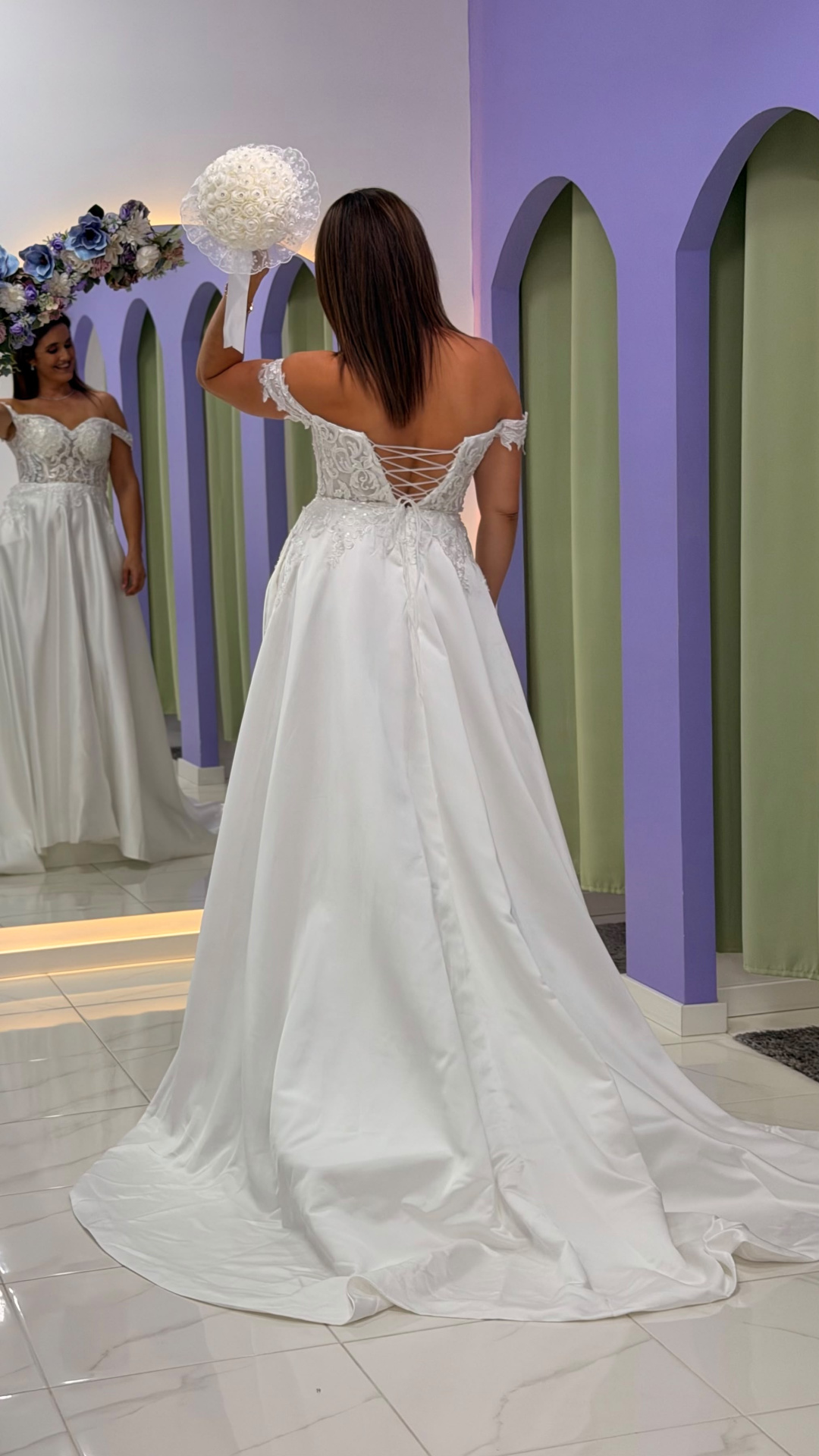 Vestido Amour Glamour Vestido de noiva branco com corpete em renda e saia longa, visto de costas, mulher segura bouquet branco em sala com parede lilás e verde e espelho.}