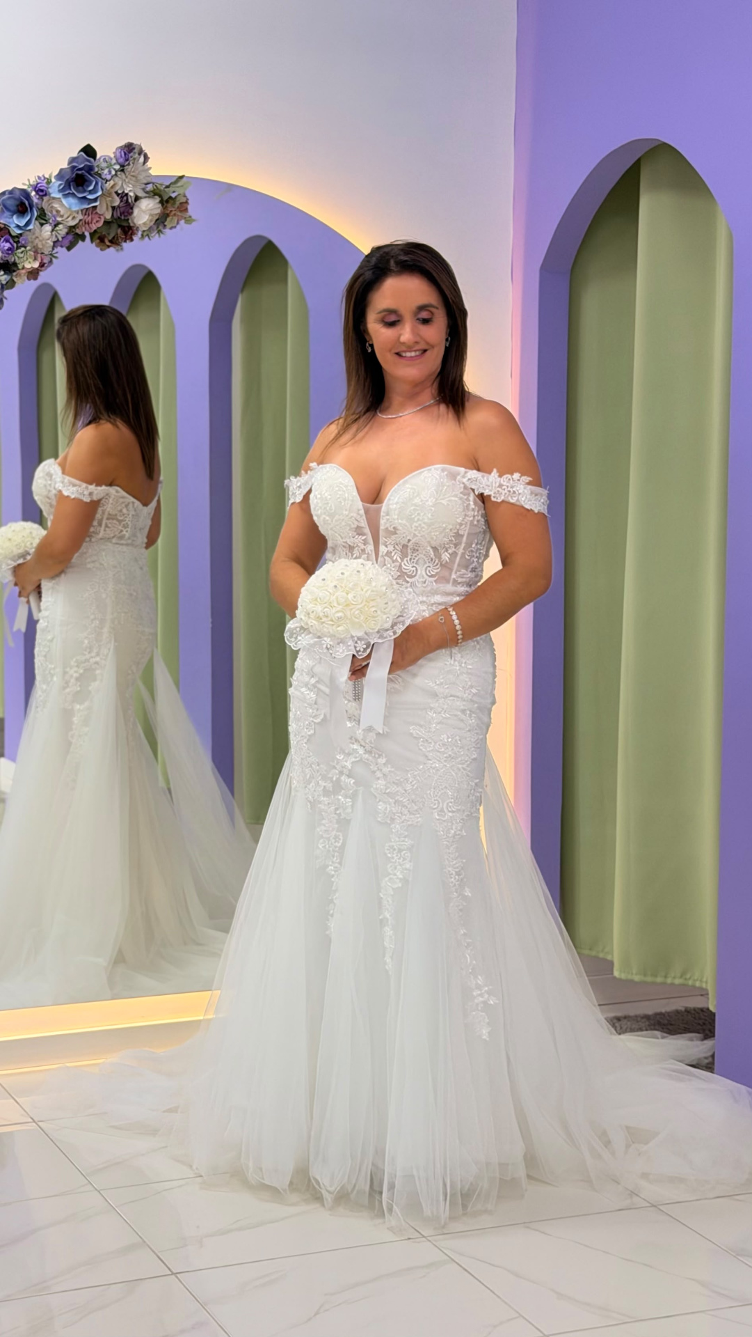 Vestido Wedding Ilusion Mulher com vestido de noiva branco e bouquet, frente a espelho com moldura lilás e cortinas verdes