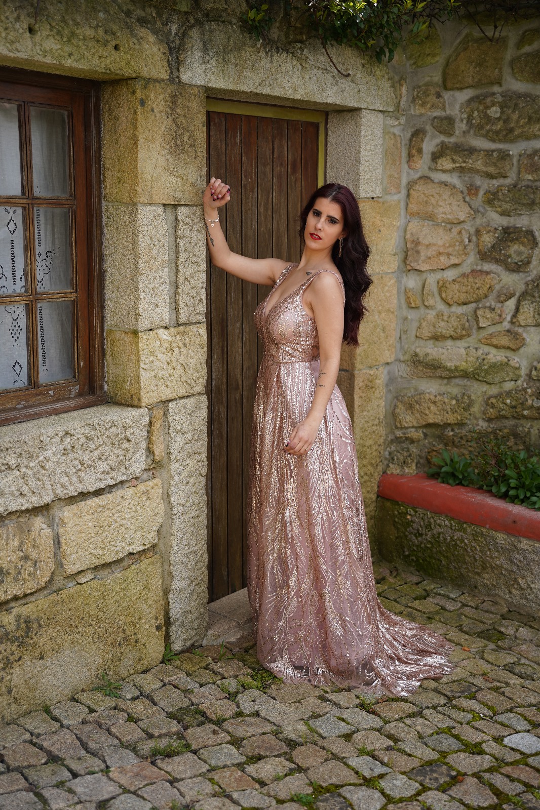 Vestido Camila Glow Mulher com vestido champanhe brilhante junto a porta de madeira e parede de pedra