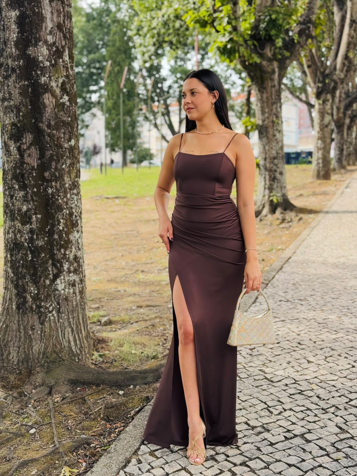 Vestido HUNGARO Vestido comprido castanho com alças finas e racha lateral, mala pequena colorida e sandálias douradas, mulher num passeio com árvores