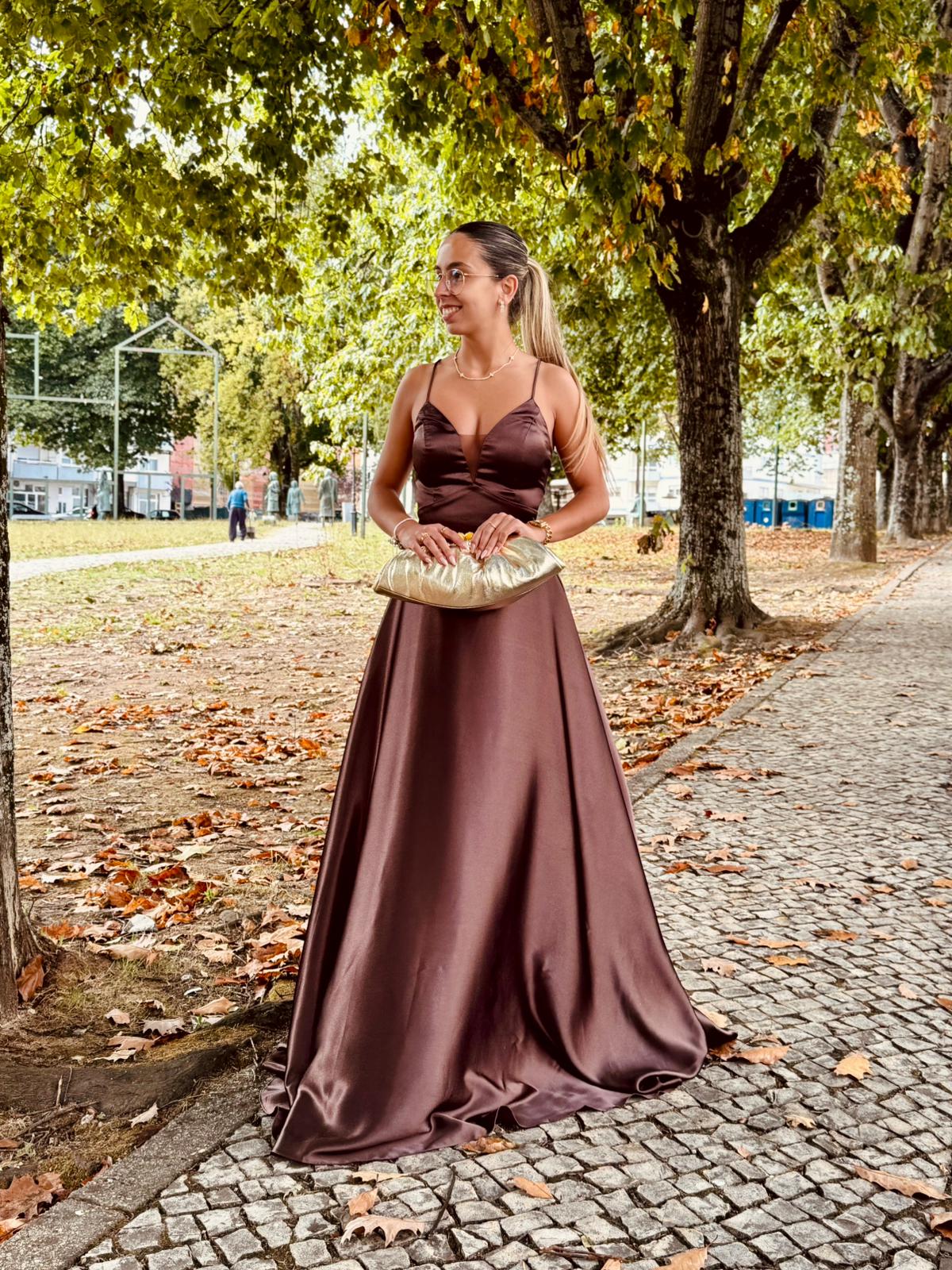 Vestido Doriane Mulher com vestido longo castanho e mala dourada num parque de árvores em outono