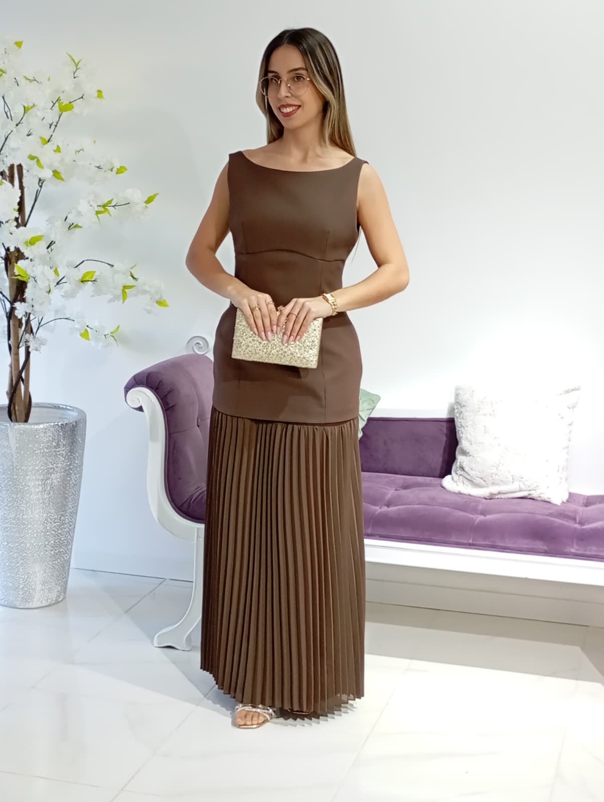Vestido Vera&Lucy Mulher com vestido castanho comprido plissado e clutch dourada ao lado de sofá roxo e vaso com flores brancas